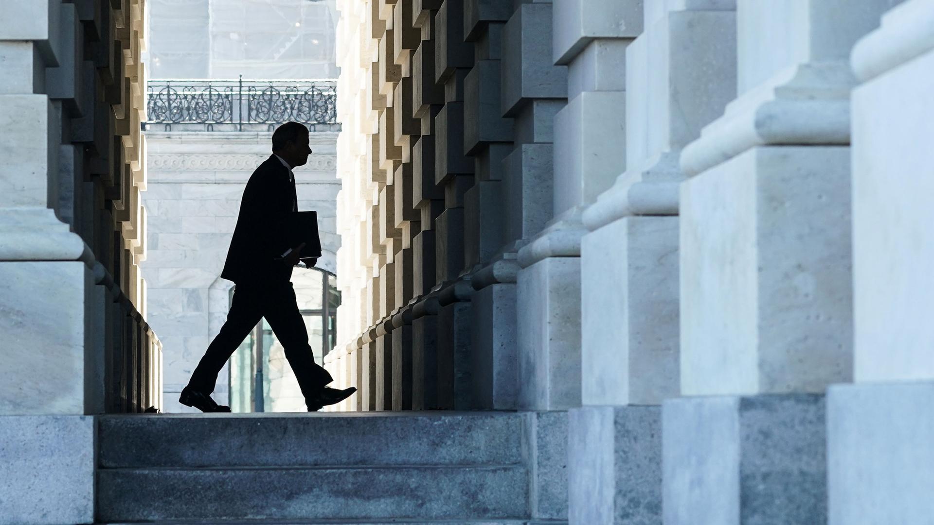 Chief Justice of the United States John Roberts is shown in shadow holding a folder and framed by the stone walls fo the Capitol building.