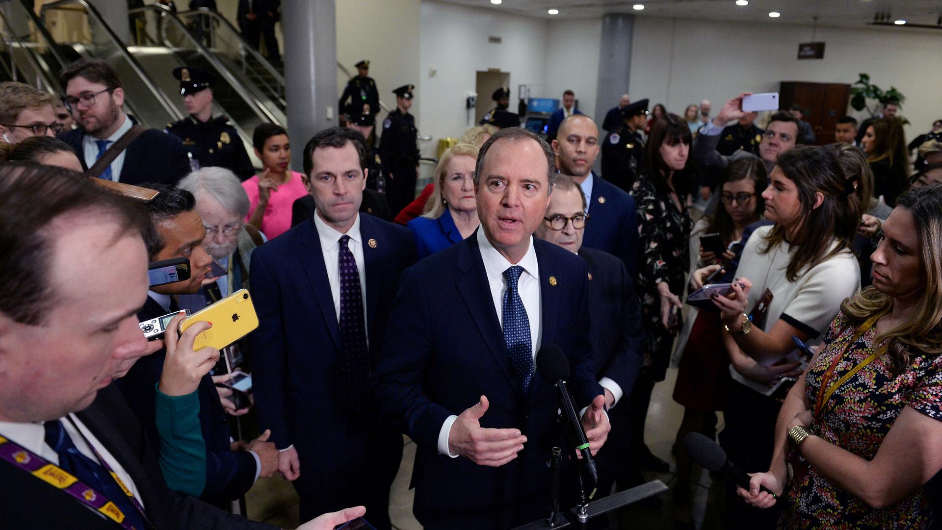 House Managers Rep. Adam Schiff is shown wearing a blue suit and tie while surrounded by reporters holding phones and other recording devices.