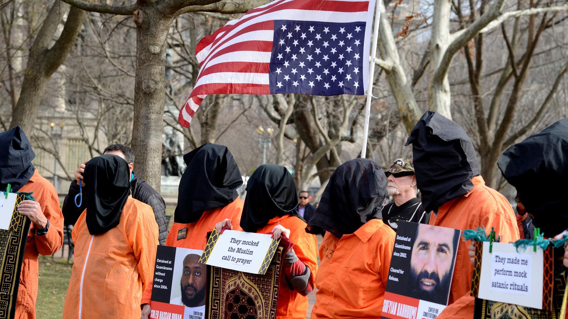 Protesters wear orange suits and black cloth over their heads with an upside down US flag behind them