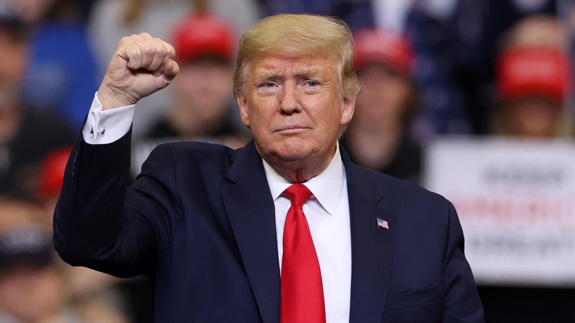 US President Donald Trump is shown wearing a blue jacket and red tie with his right hand raised in a fist.