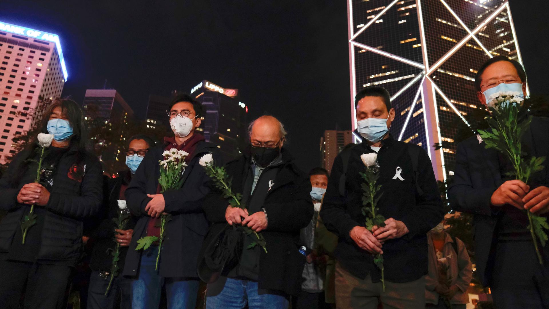 A group of people are shown holding flowers and wearing face masks with skyscrapers in the distance.