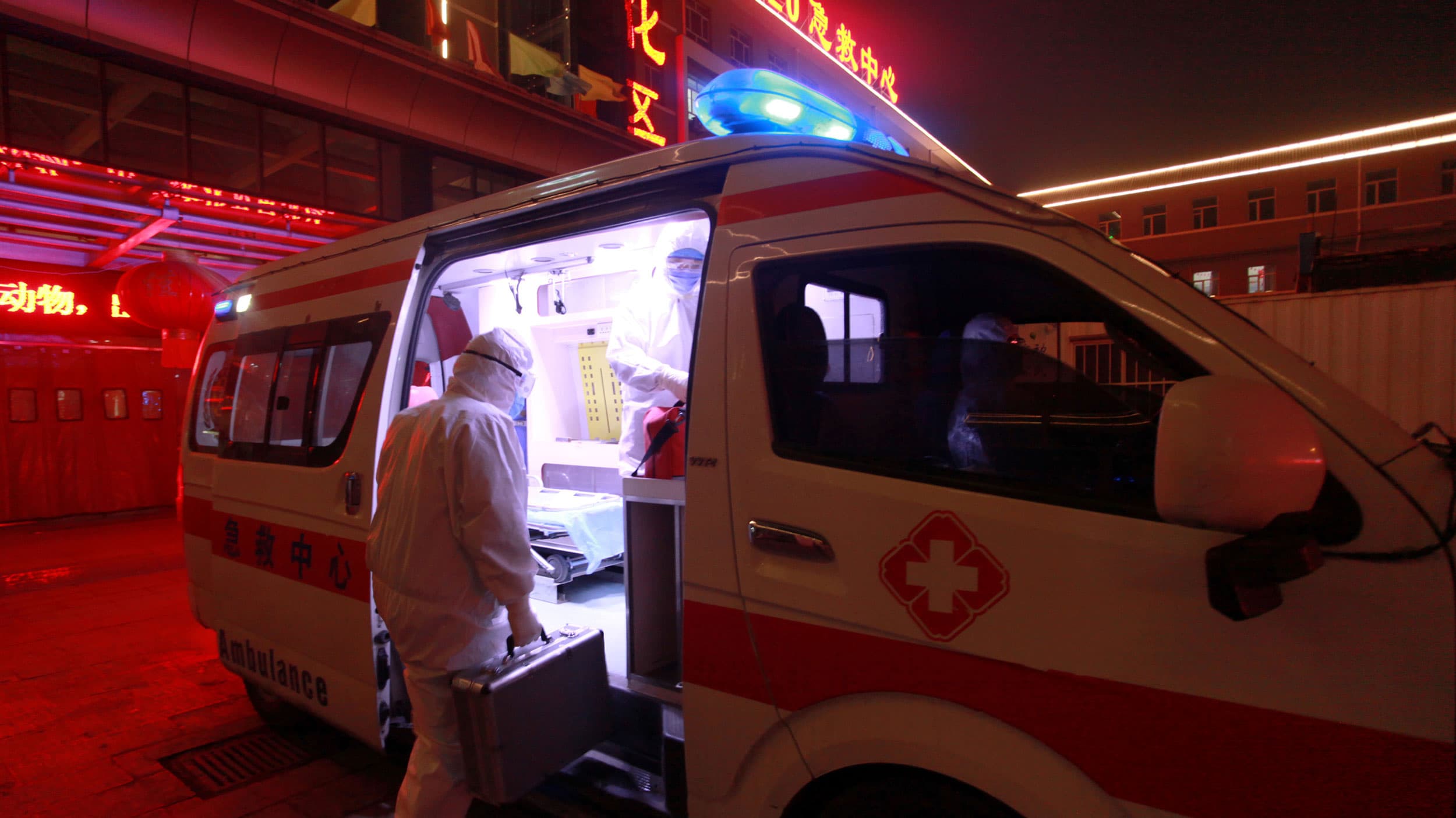 An ambulance worker wearing a full body protective suit is shown carrying a suitcase and climbing into an ambulance van.