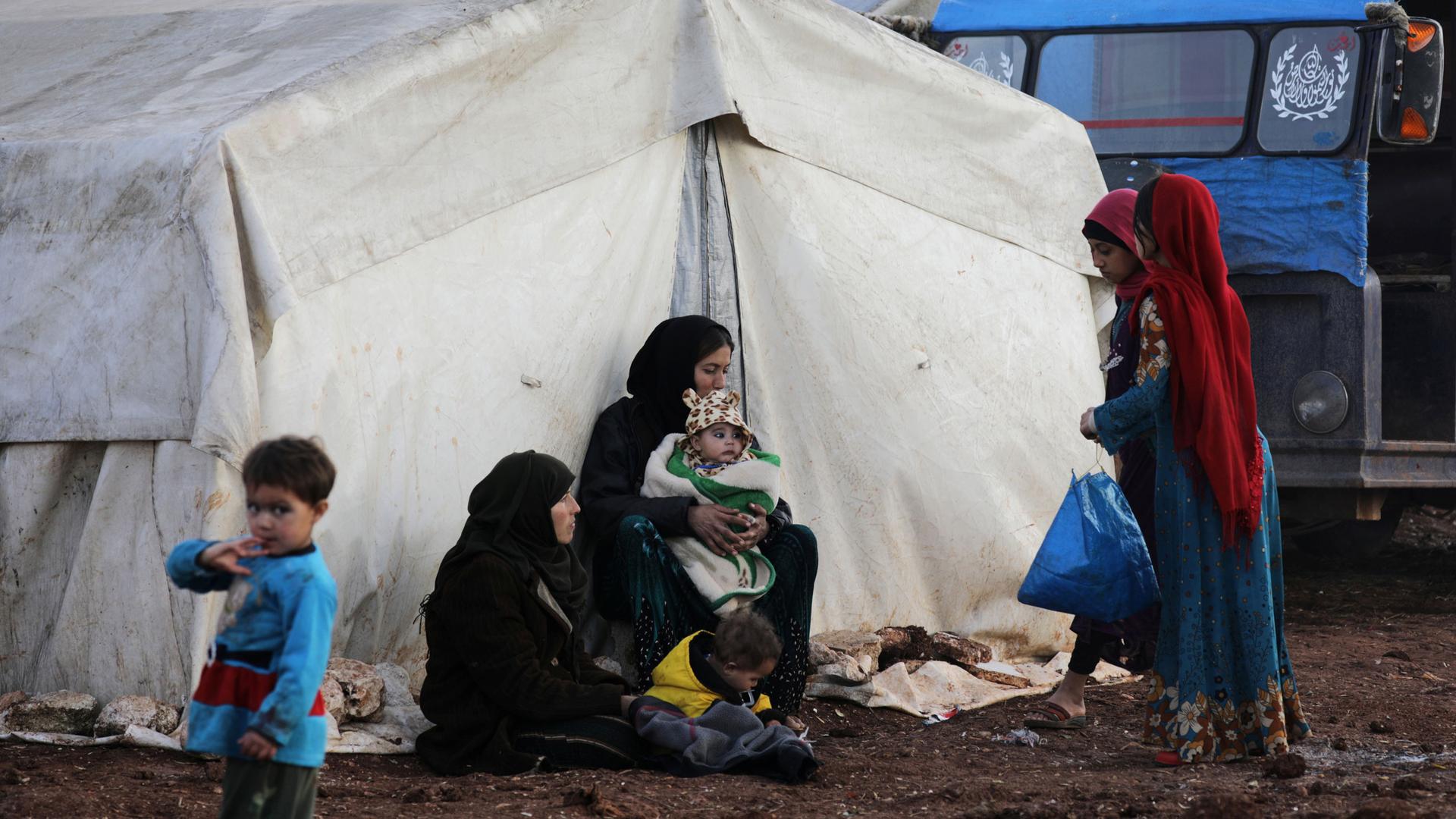 Two women wearing black hijabs are shown with several children in front of a white tent.