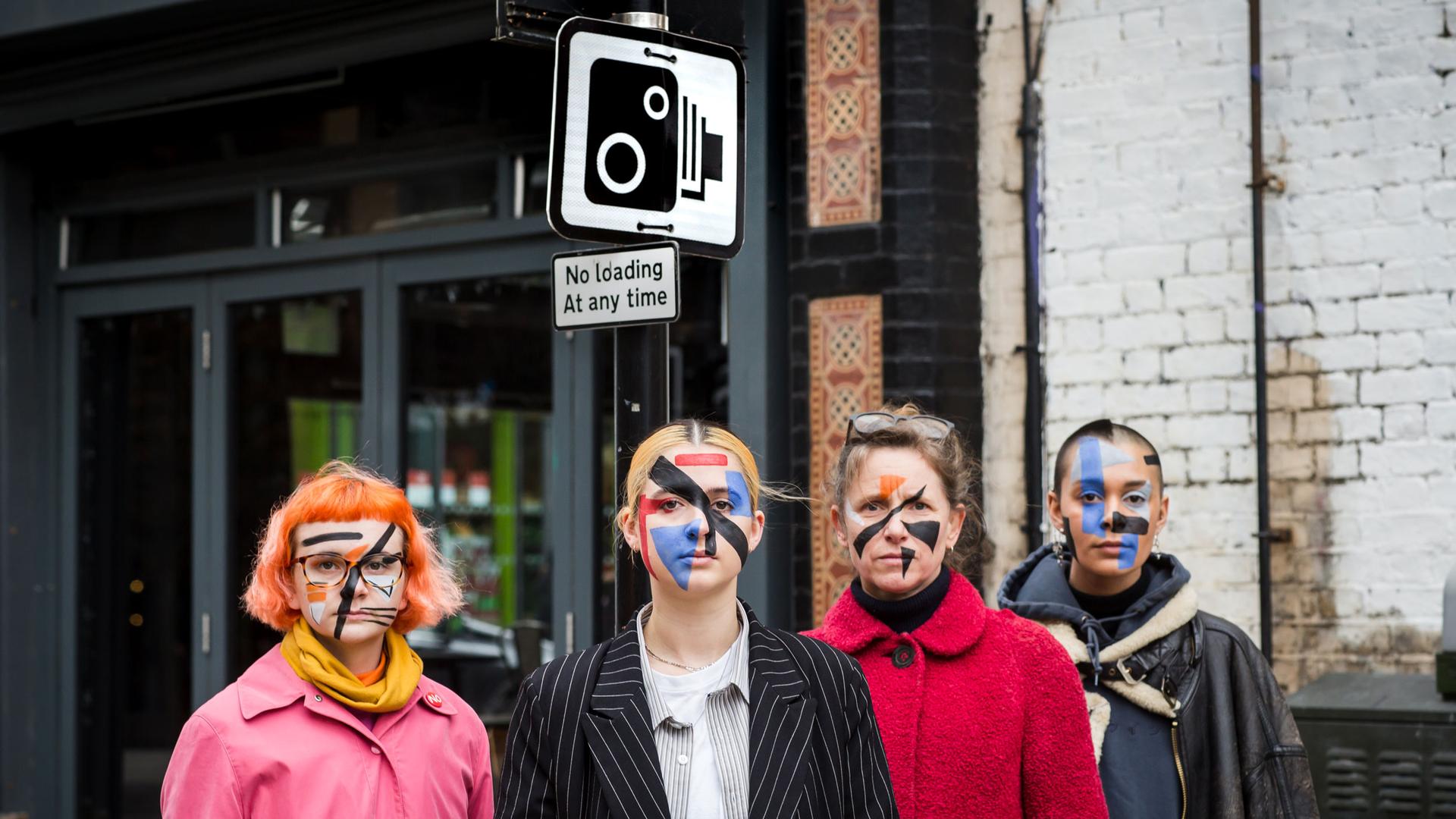 Four people with painted faces stand in front of a street sign with a camera on it.