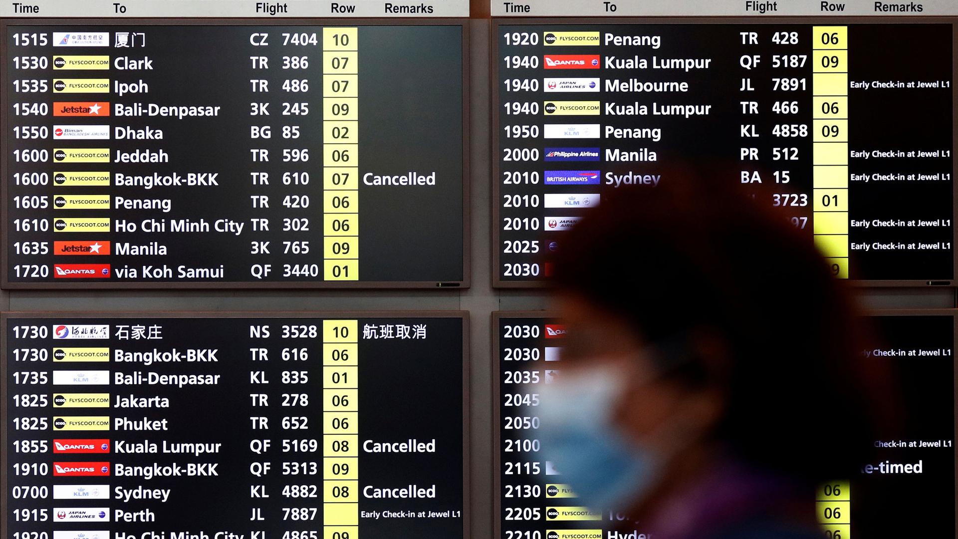 A woman wearing a mask passes is shown in soft focus with a arrivals and departures flight information board behind her.