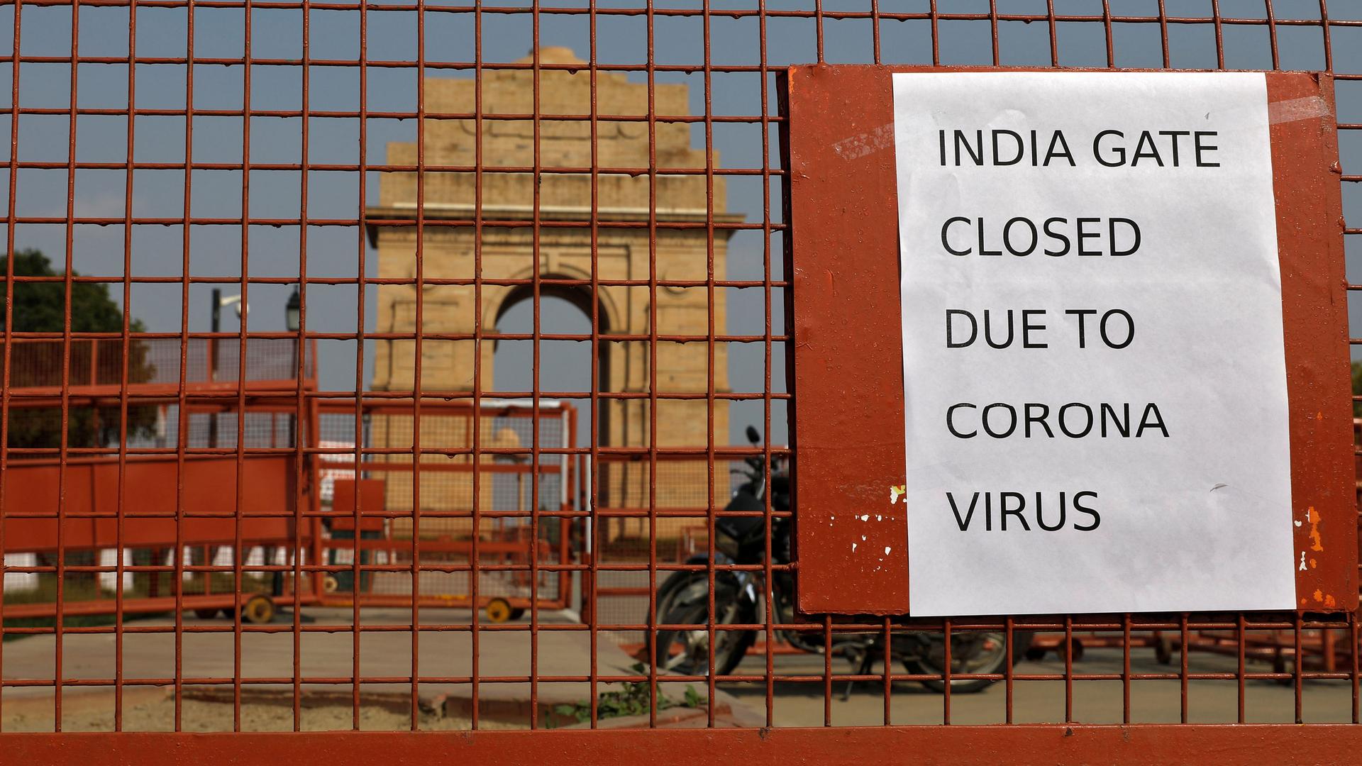 A close-up photograph of a reddish metal gate with a motorcycle parked behind it and the India Gate war memorial is shown off in the distance.