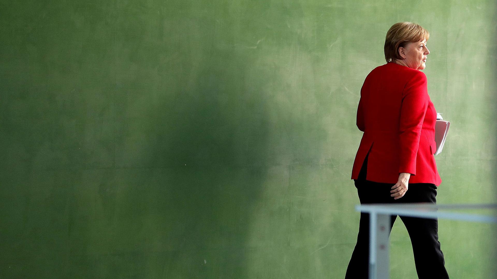 German Chancellor Angela Merkel is shown walking next to a green wall and wearing a red suit jacket.