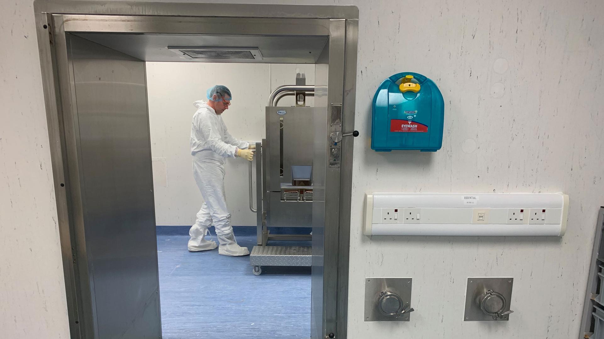 A scientist is shown through a doorway wearing protective medical clothing while working at a machine.