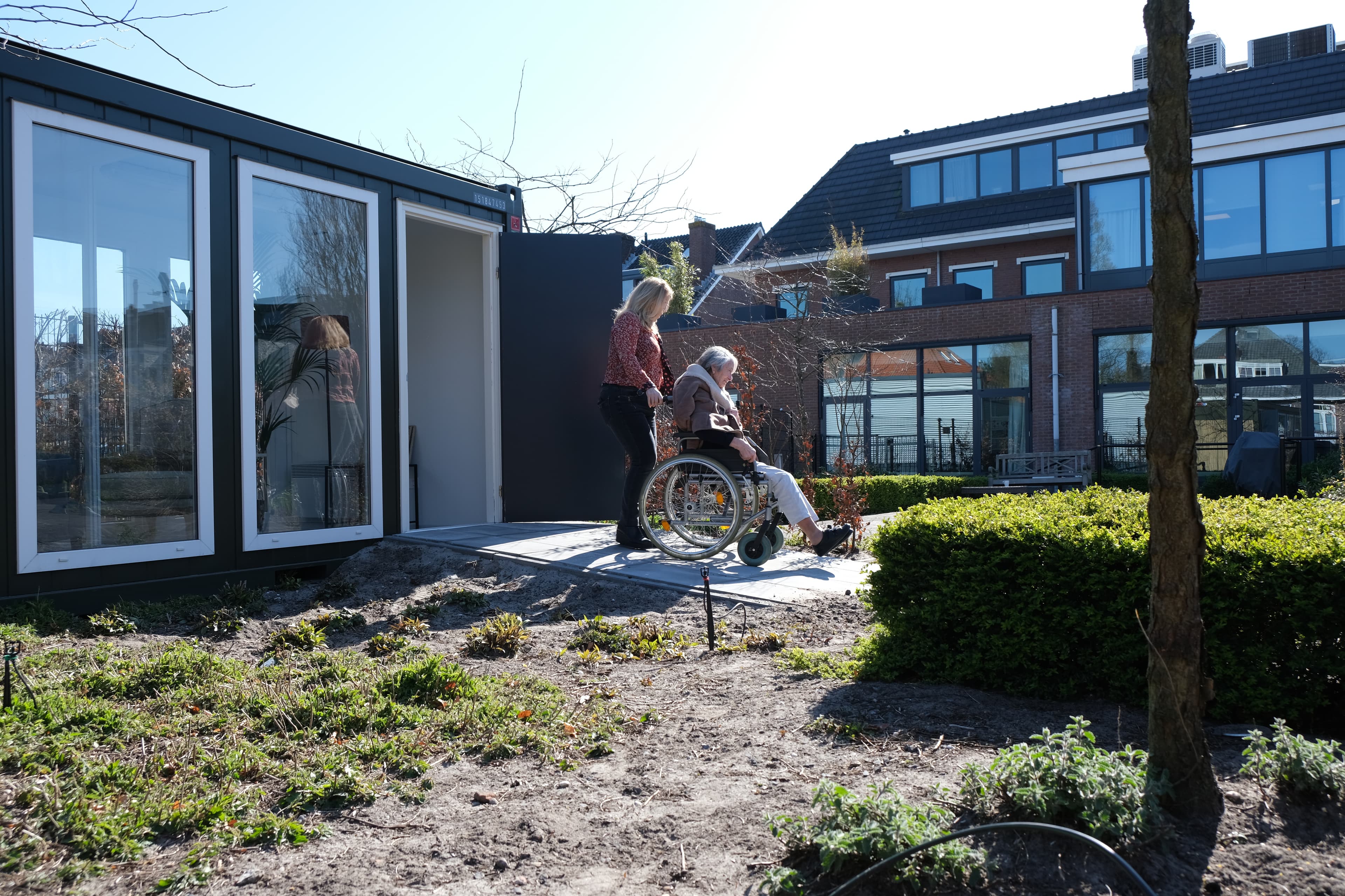 A woman pushes another woman in a wheelchair outside a glass garden house.