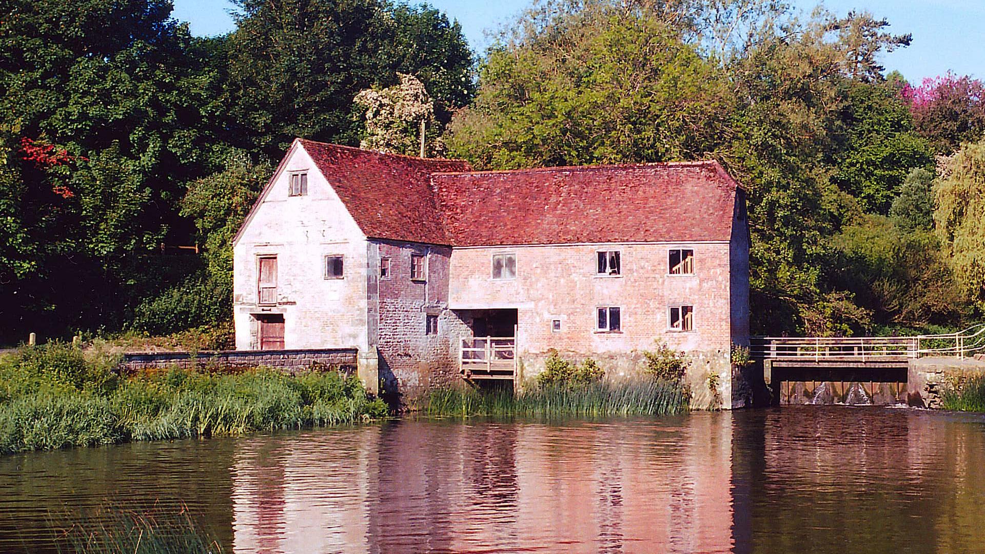 The Sturminster Newton Mill brick and stone building is shown across a body of water with its refection in the water.