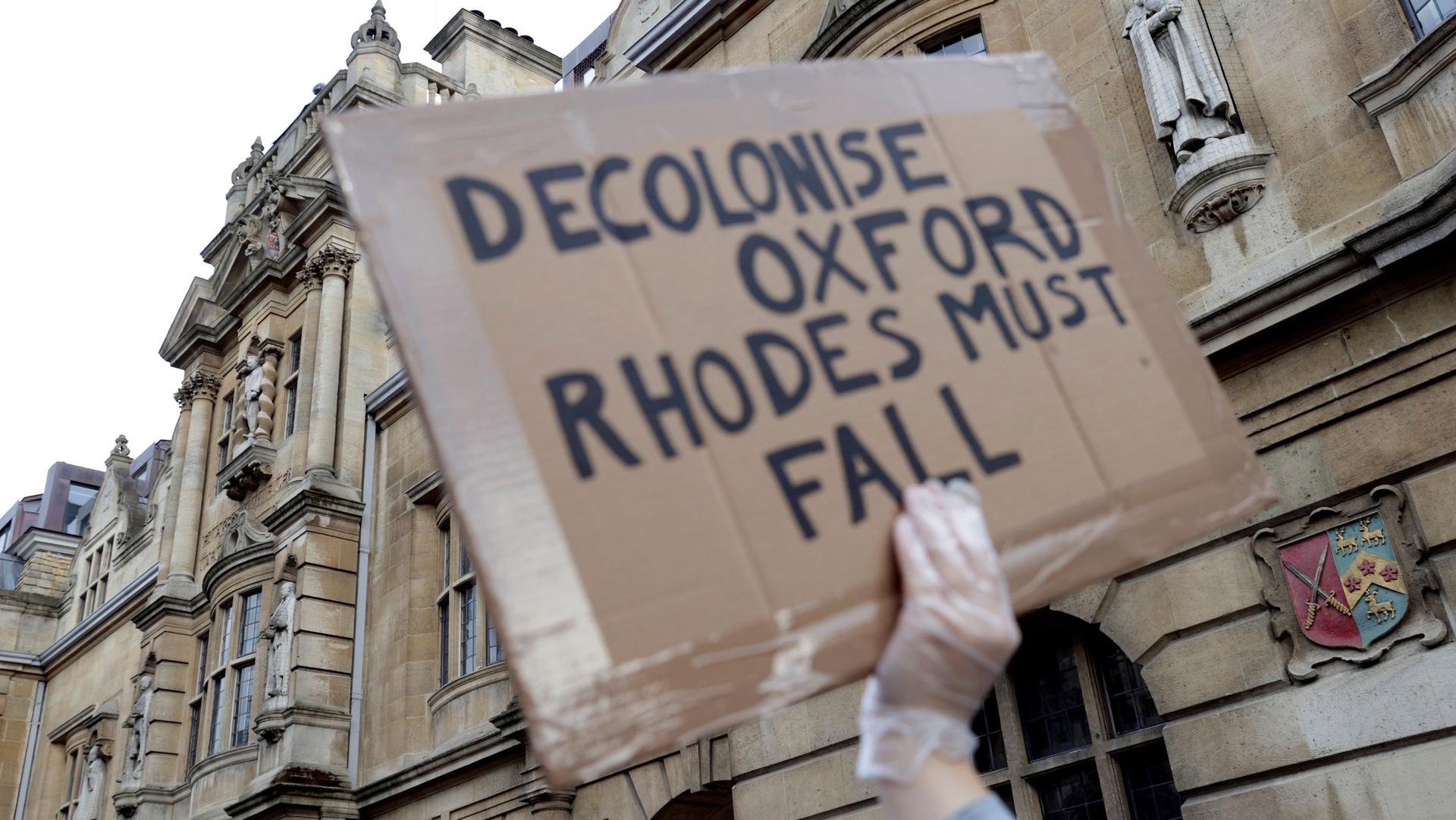 A demonstrator's hand is seen holding a placard reading "Decolonise Oxford/Rhodes must fall"
