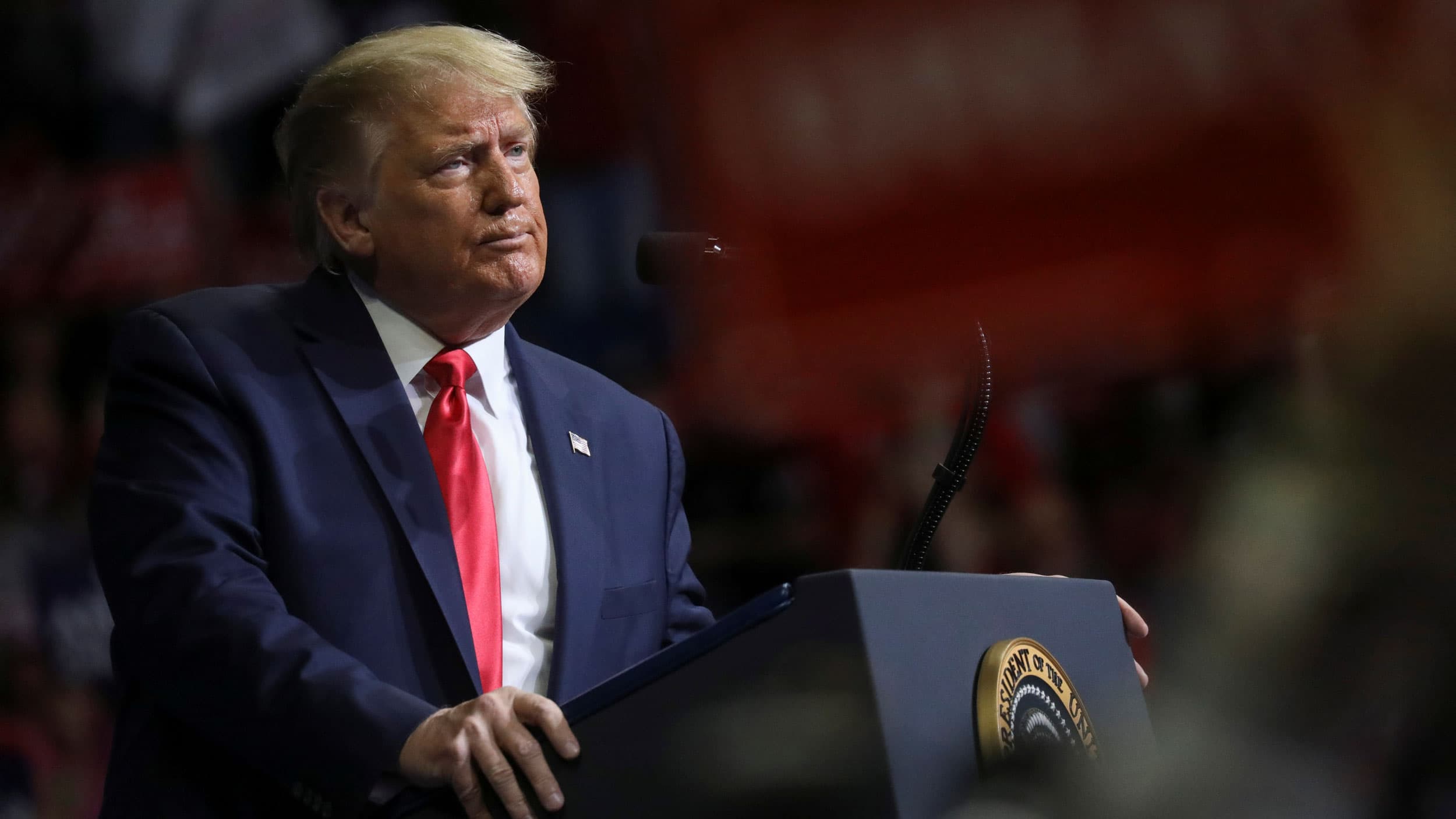 Donald Trump is show with his hands on either side of a podium while wearing a dark suit and red tie.