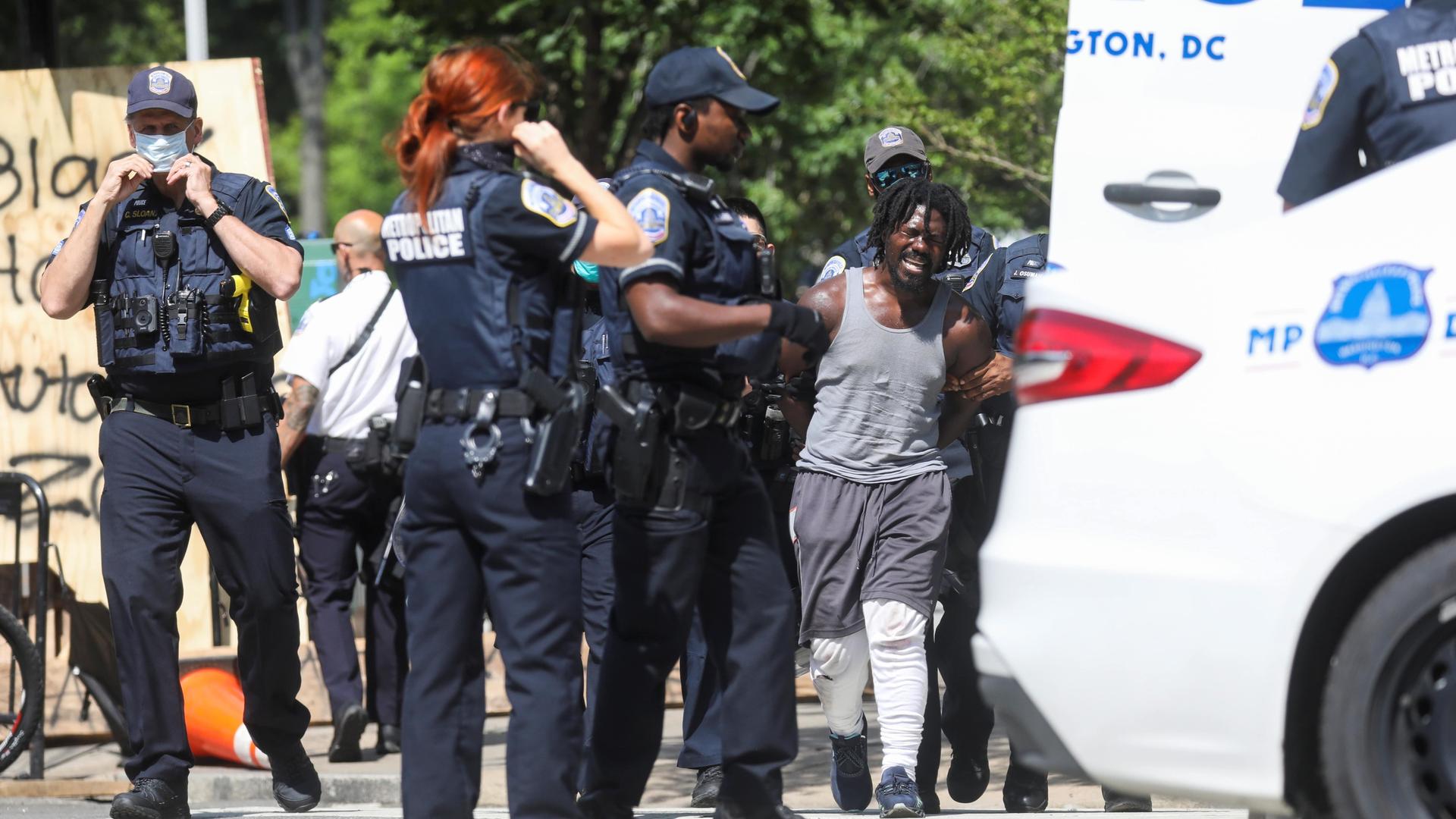 A group of police officers in black uniforms detain a Black man wearing gray pants and white tank.
