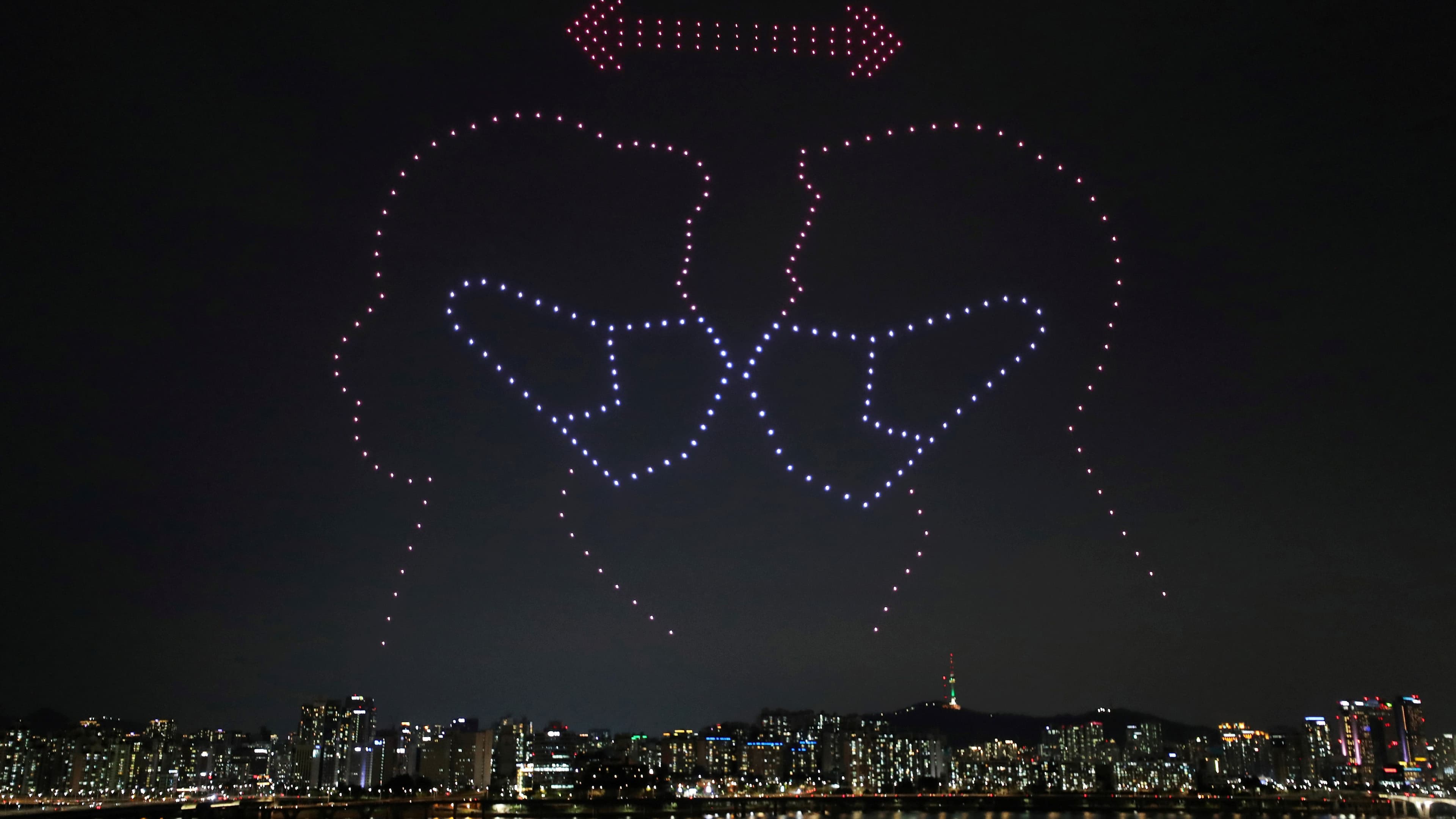 A silhouette of 2 people wearing a mask appears over the night sky of a city.