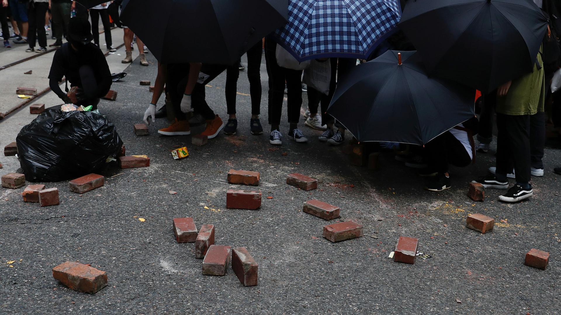 A crowd of people hold umbrellas toward the camera with several bricks laying in the street.