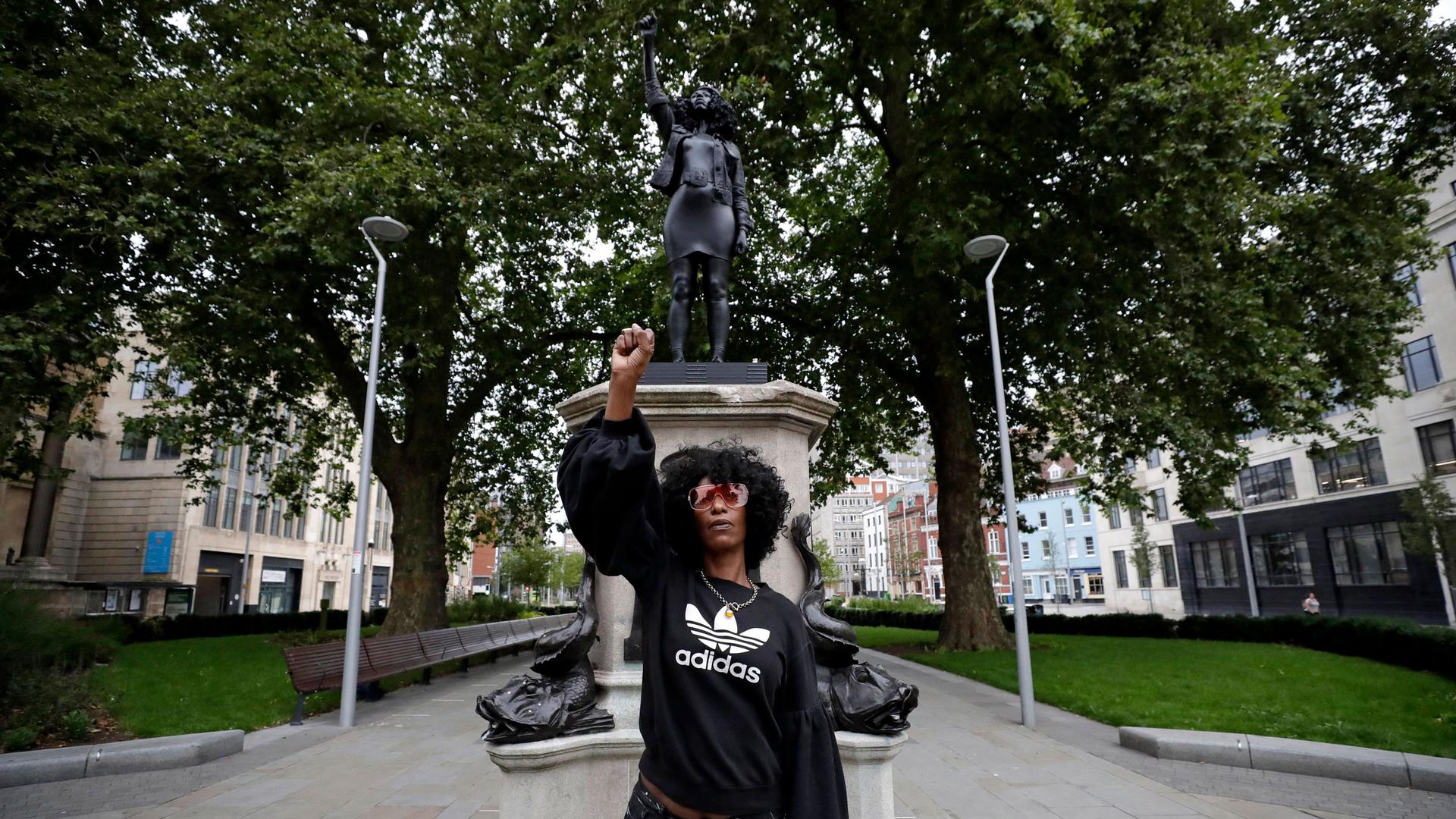 A woman wearing a dark jacket and sunglasses is shown standing in front of a statue of her with her hand raised.