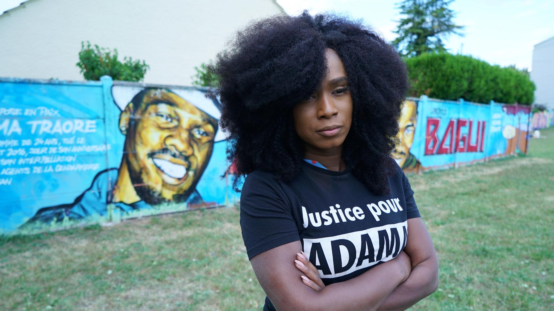 A black woman wearing a black shirt with white lettering poses near a mural