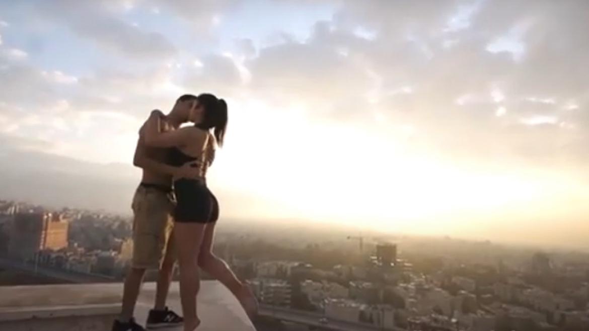 A man kisses a woman wearing blach shorts on a rooftop overlooking a pink skyline