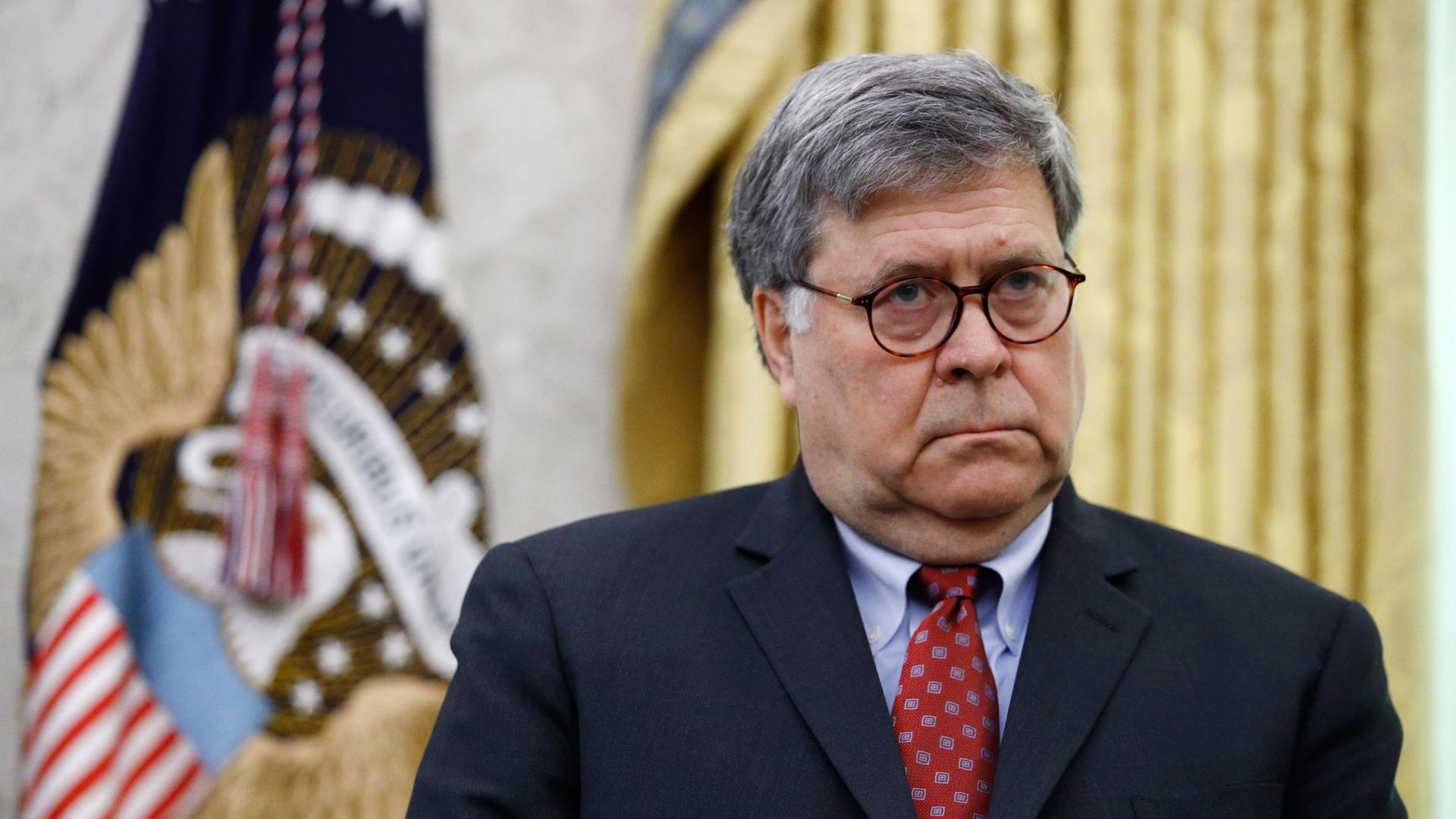 Attorney General William Barr is shown wearing a dark suit and red tie while standing in front of the POTUS flag.