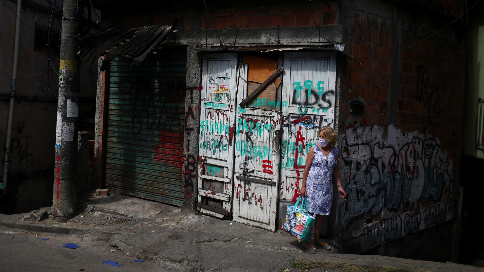 A woman is shown wearing a medical mask and carrying a bag while standing in front of doors with graffiti painted on them