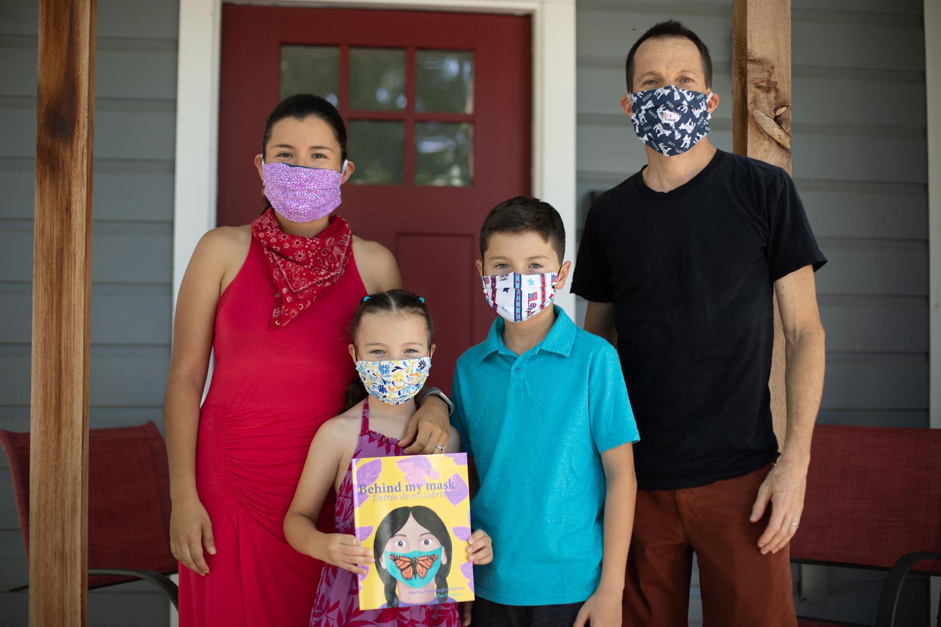 A family of four pose for a photo wearing masks. The young girl holds a book in her hands.