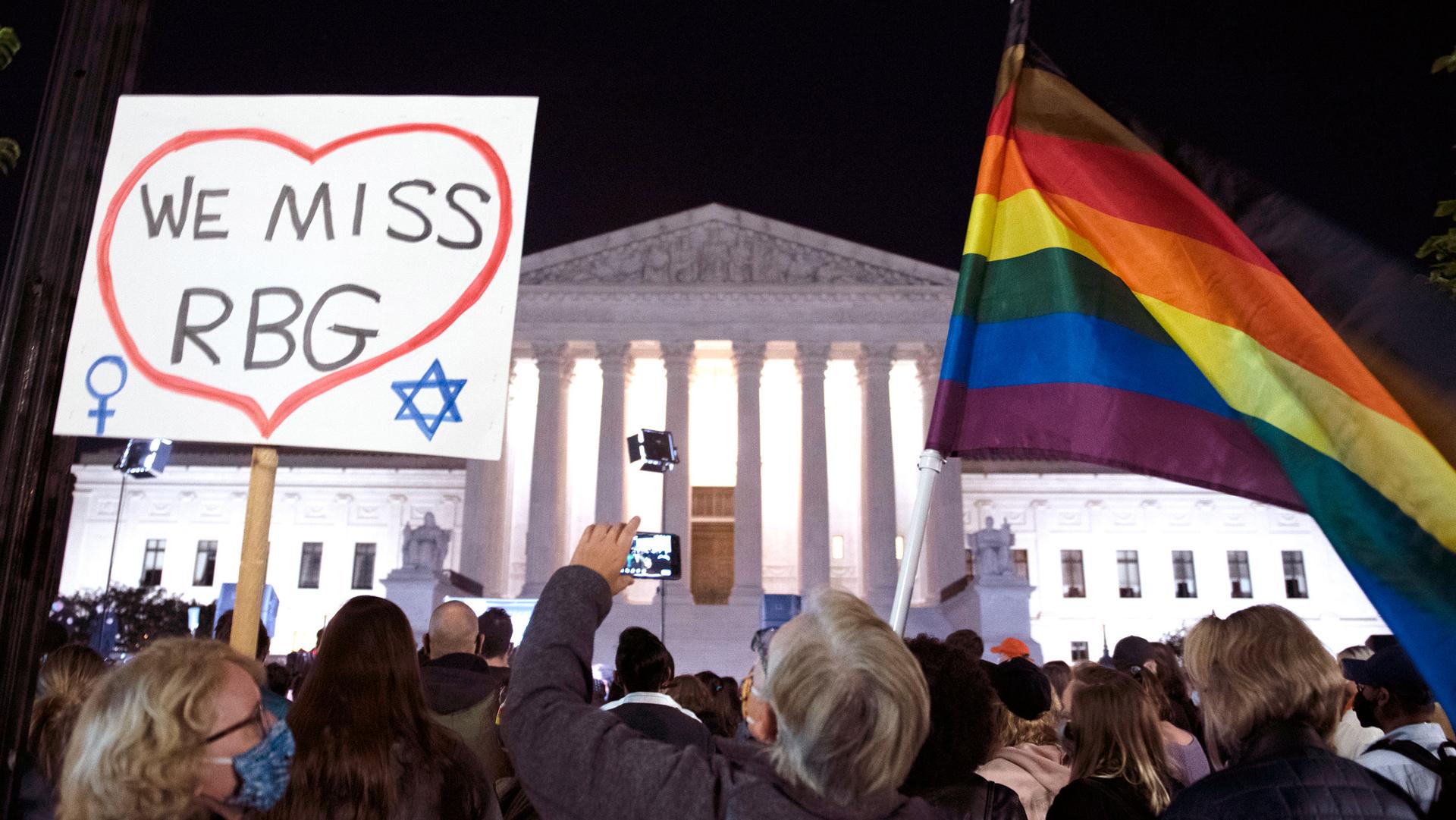 A crowd of people are shown in the dark of night outside of the brightly lit facade of the US Supreme Court and holding signs and flags.