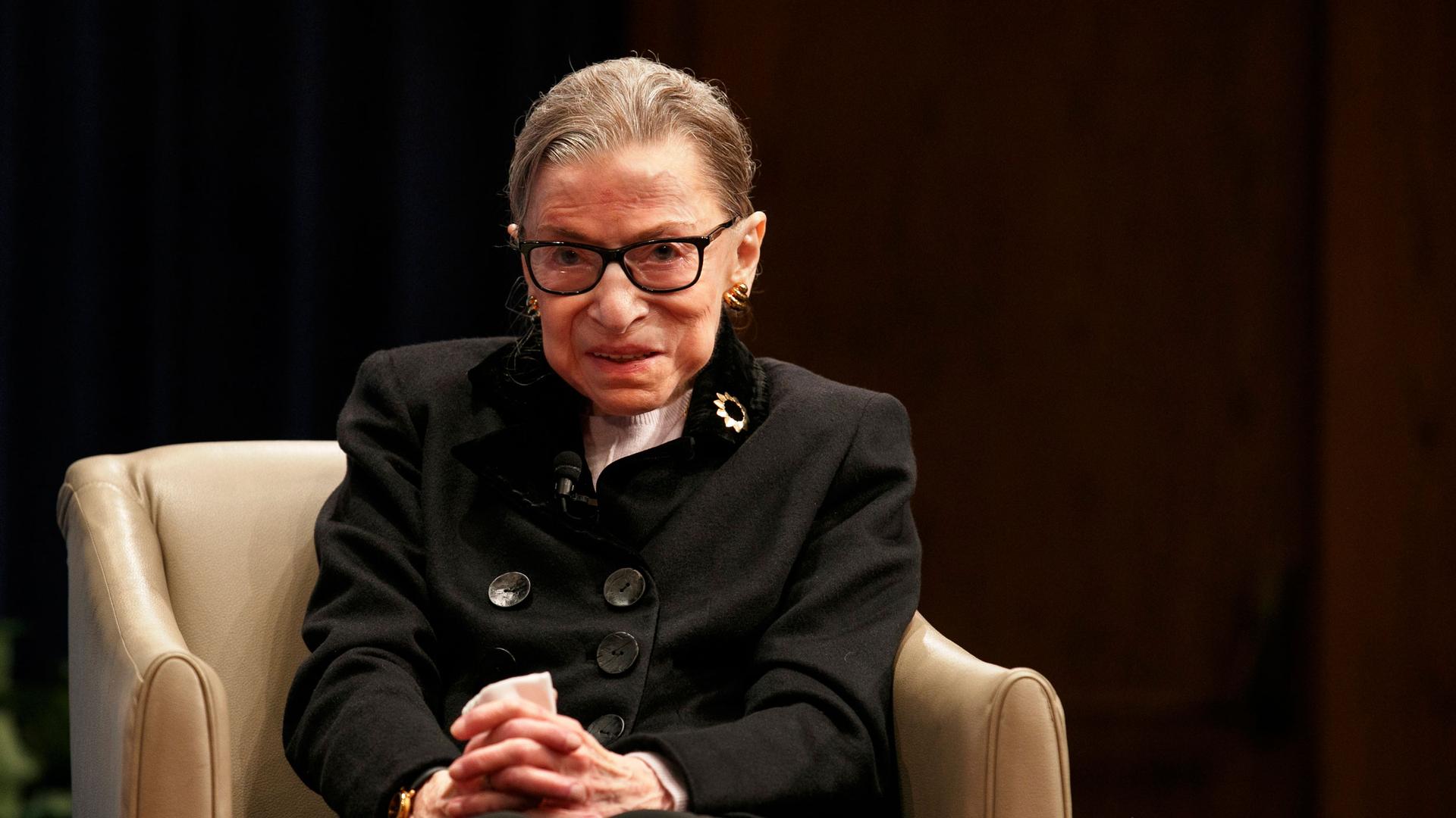 Supreme Court Justice Ruth Bader Ginsburg is shown sitting in a tan-color armchair and wearing a dark jacket with large buttons on the front.