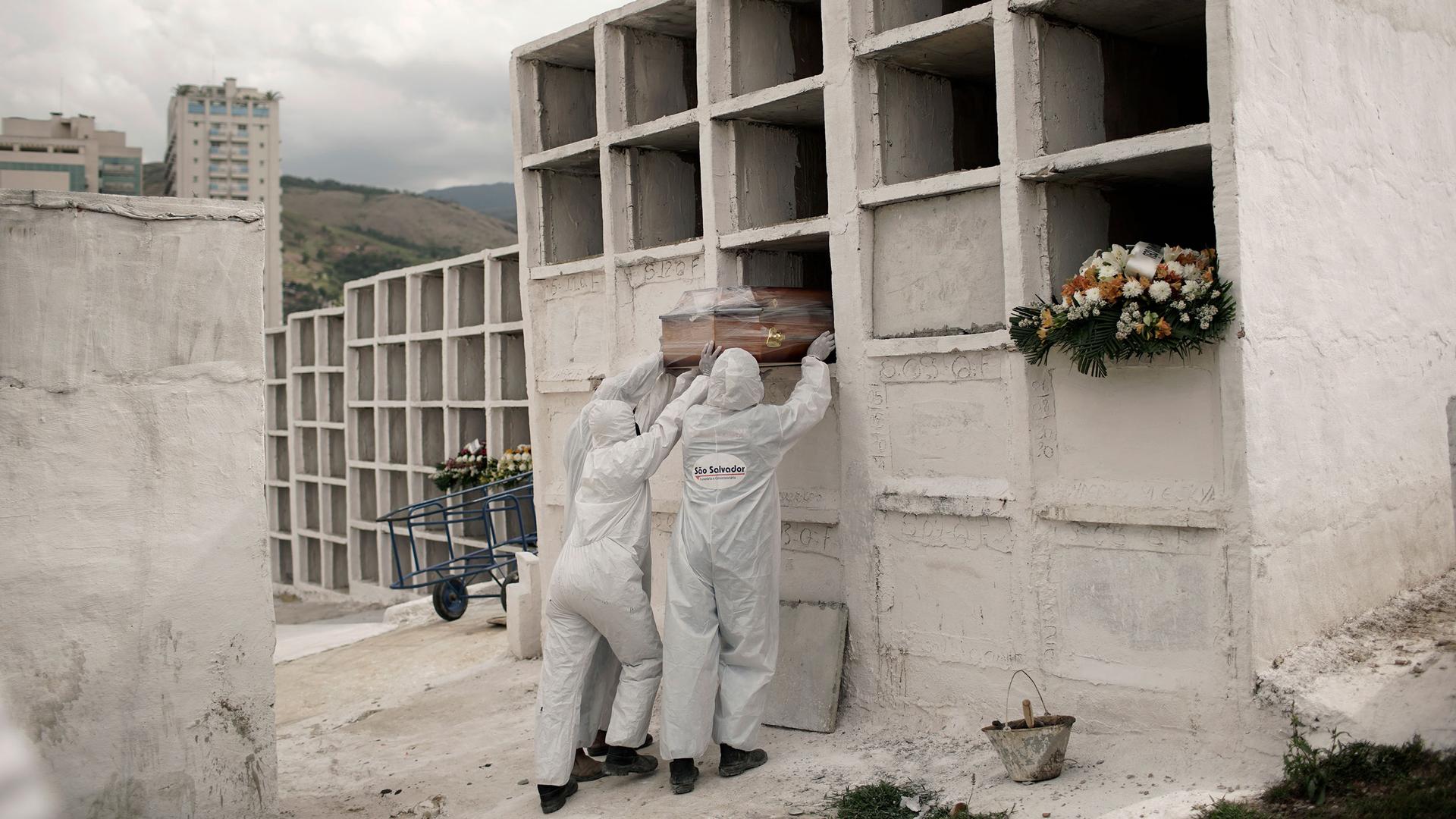 Three workers are shown wearing white protective clothing and placing a coffin into an empty slot in large white niche.