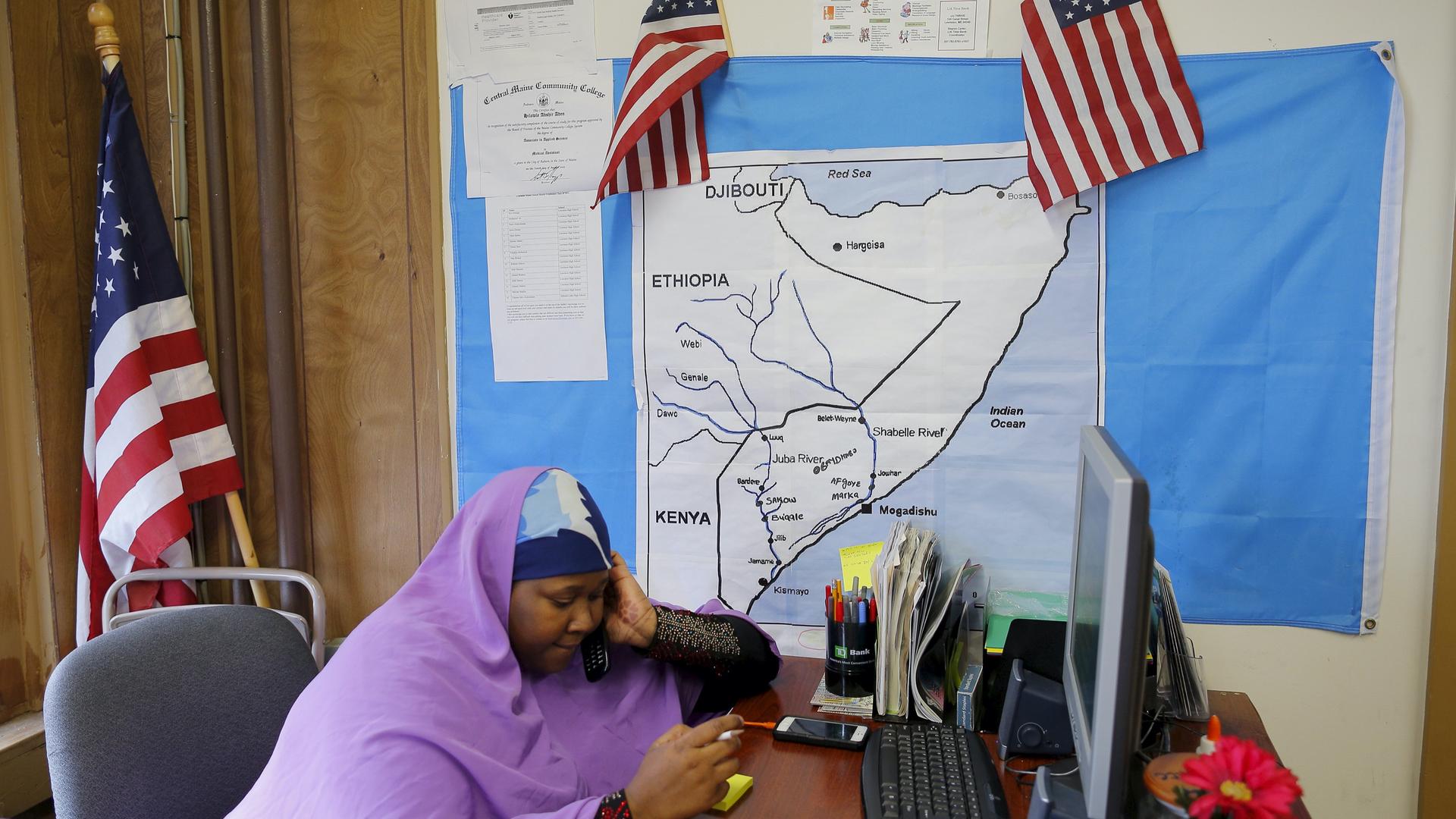 A Somali woman wearing a purple headscarf answers the phone in an office with American flags and East Africa map on the wall.