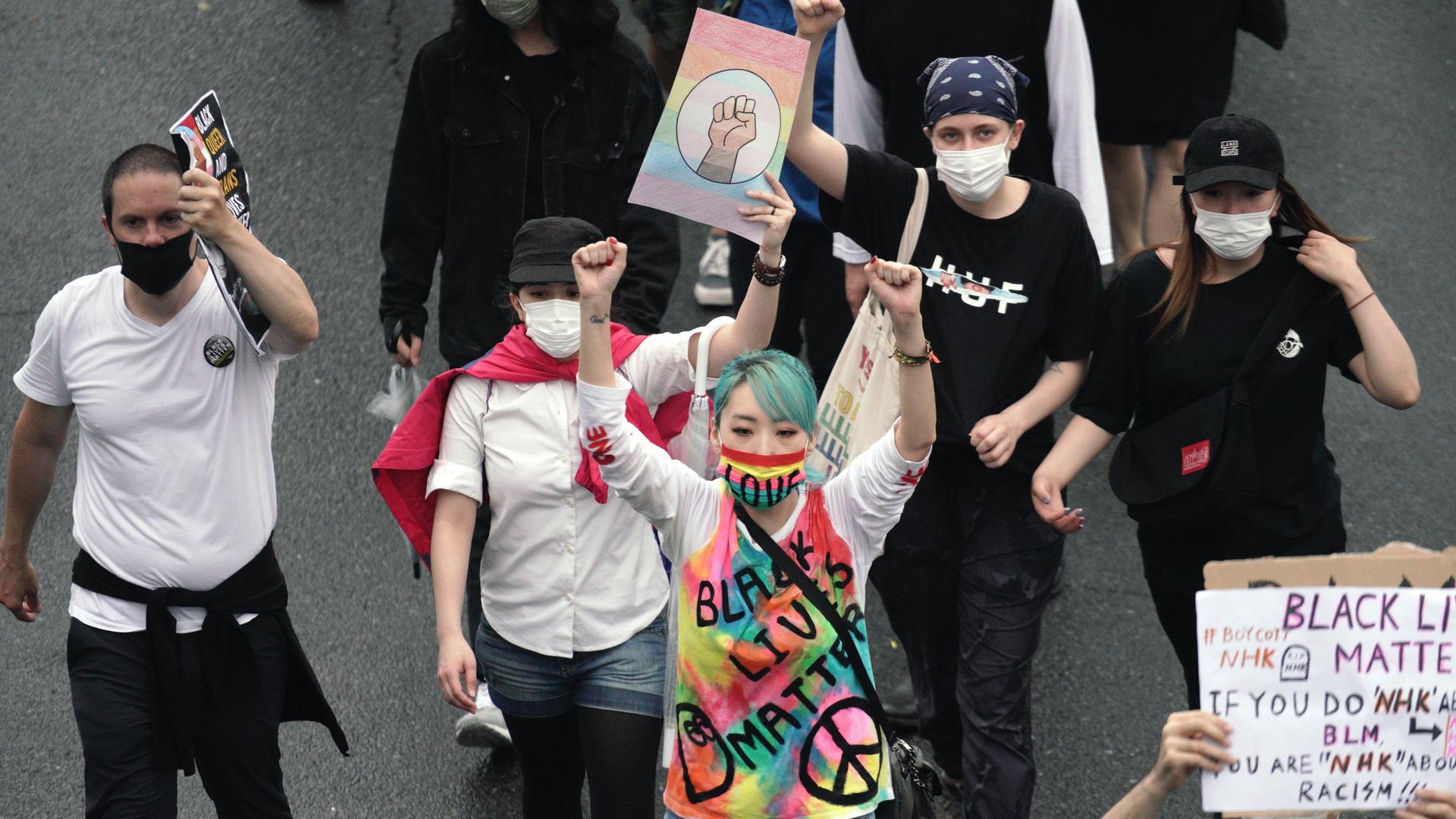 A group of protesters carries protest signs. One wears a colorful tie-dye shirt and has blue hair. The others wear white and black shirts.