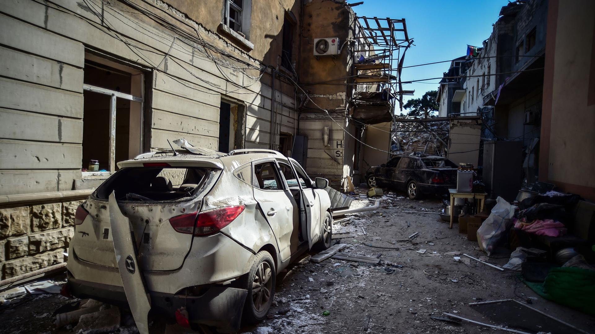 A heavily damaged car is shown in an alley in the nearground with several damaged buildings in the distance.