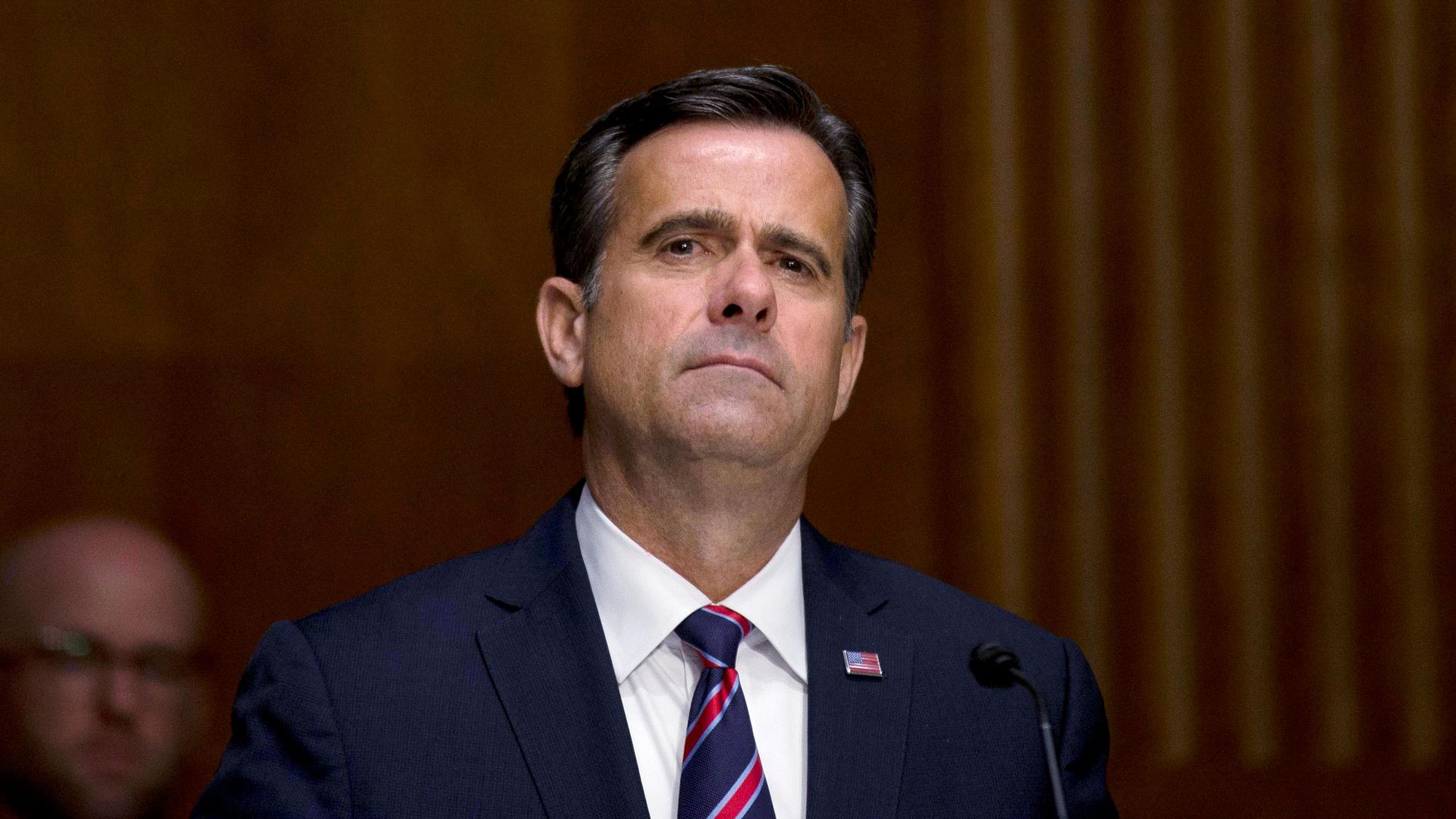 Director of National Intelligence John Ratcliffe is shown wearing a dark blue suit and striped tie with a US flag pin.