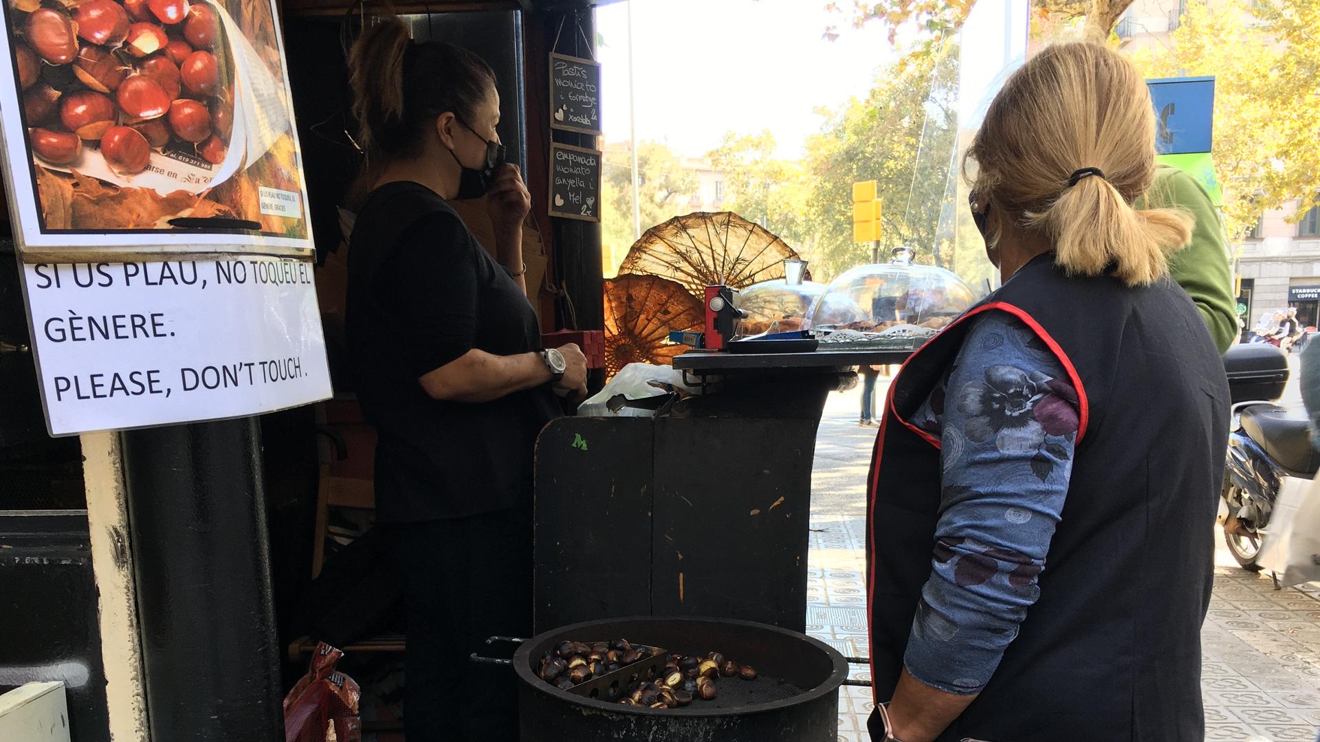 A woman wearing a face mask sells roasted chestnuts as another woman wearing a blue apron minds the chestnuts.