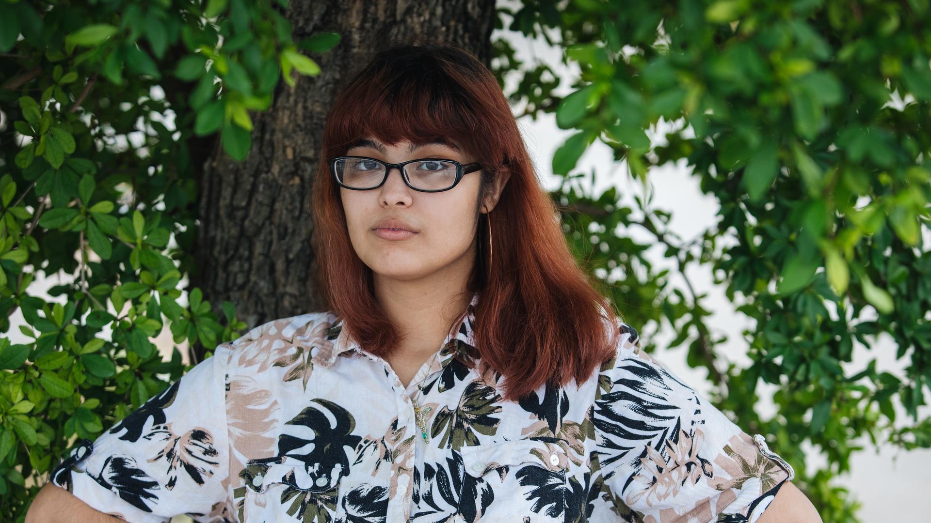 A young person with glasses and wearing a floral shirt poses under a tree outside