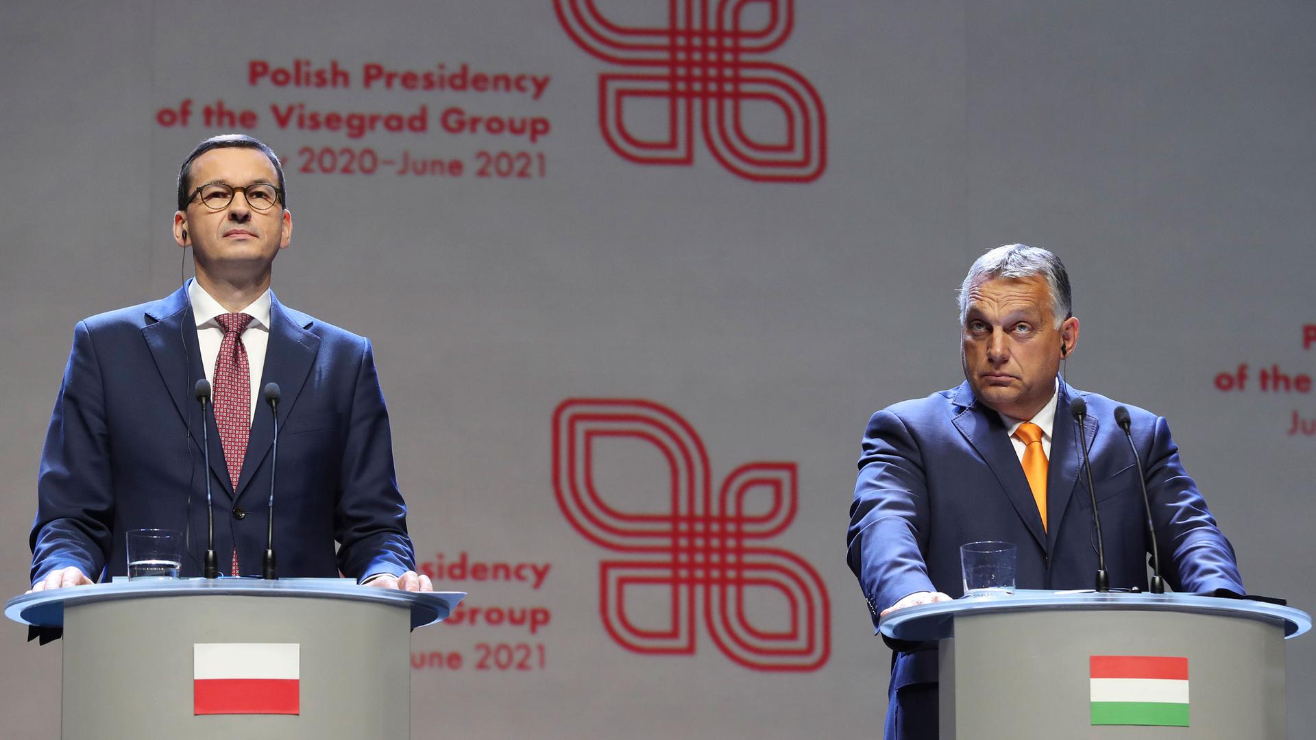 Viktor Orban (R) prime minister of Hungary and Polish Prime Minister Mateusz Morawiecki are shown both wearing dark suits and standing behind podiums, each with their respective country flags on the front.