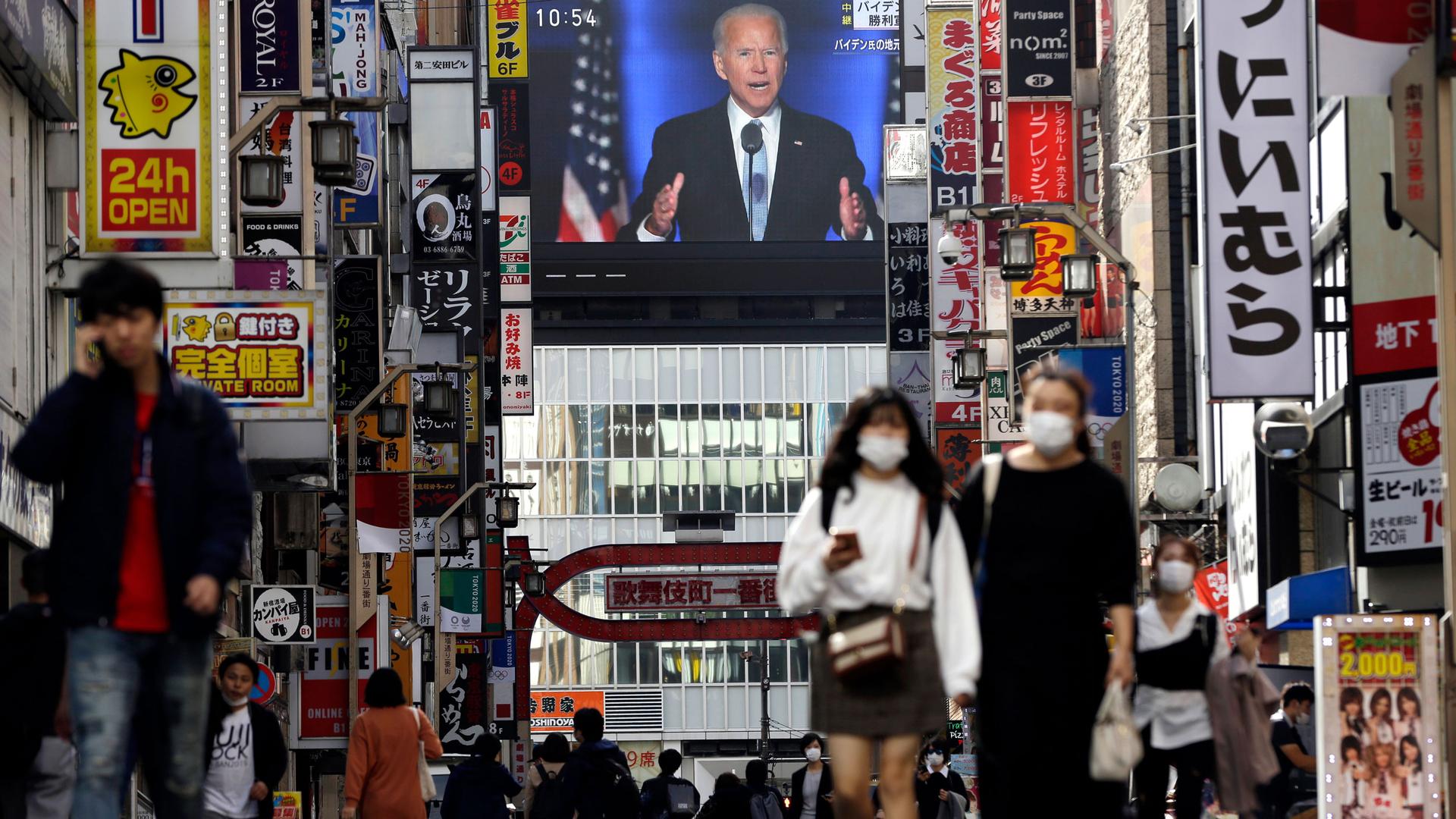 The busy streets of the Shinjuku shopping district in Tokyo adorned with signs for stores and restaurants lining either side while a live broadcast shown on a large public television monitor of President-elect Joe Biden.