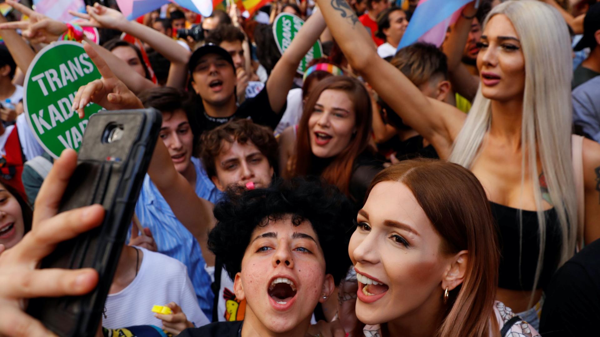 Several people surrounded by rainbow flags in a crowd cheer together in a parade and two people in front take a selfie