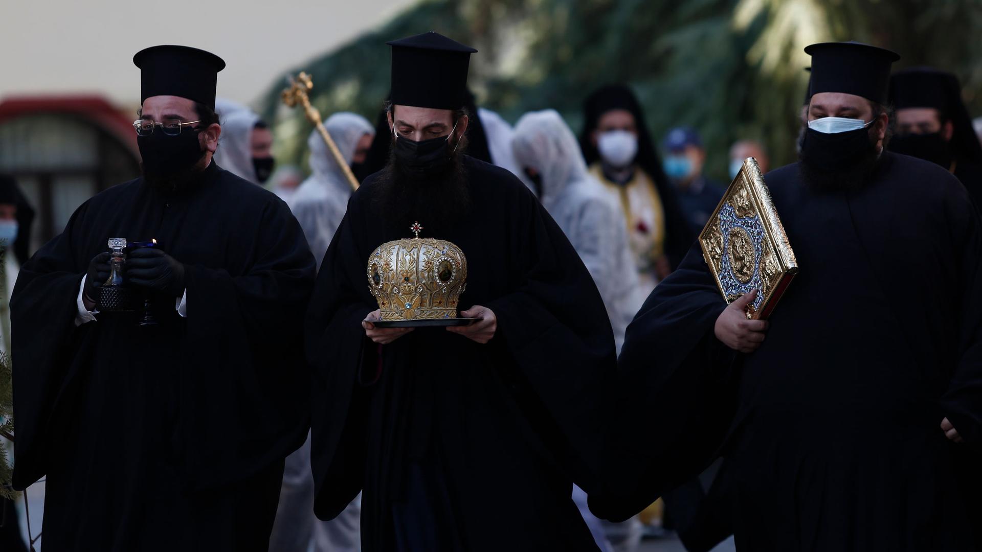 Three priests are shown wearing face masks and dressed in traditional black Greek Orthodox attire while carrying religious objects.