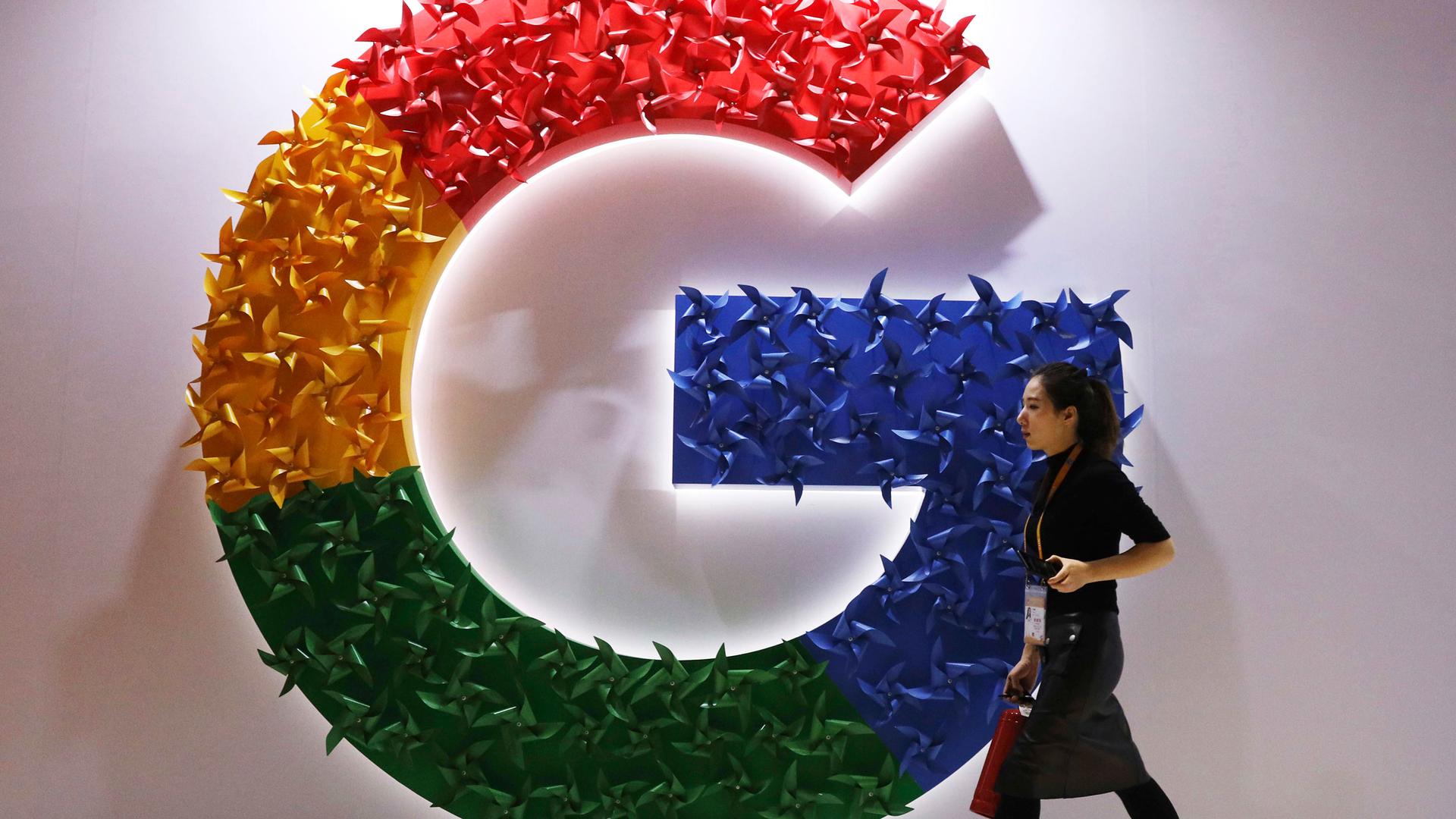 A woman wearing a skirt and name badge walks past the logo for Google affixed to the wall.
