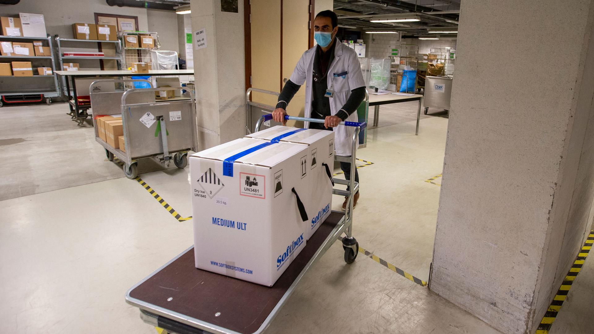 A man is shown wearing a white medical jacket and pushing a cart with two large white boxes on top sealed with blue tape.