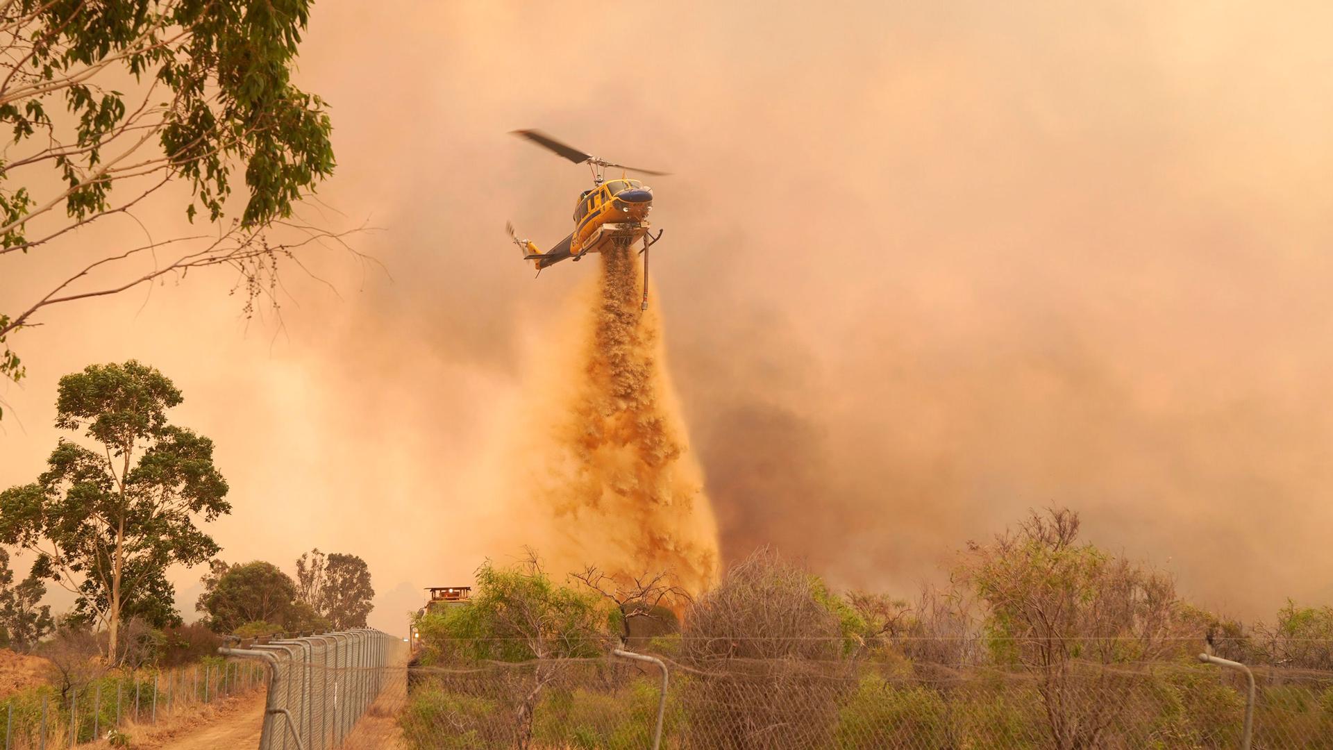 A helicopter is shown flying above a wooded area with a large metal fence on one side and dropping fire retardant.