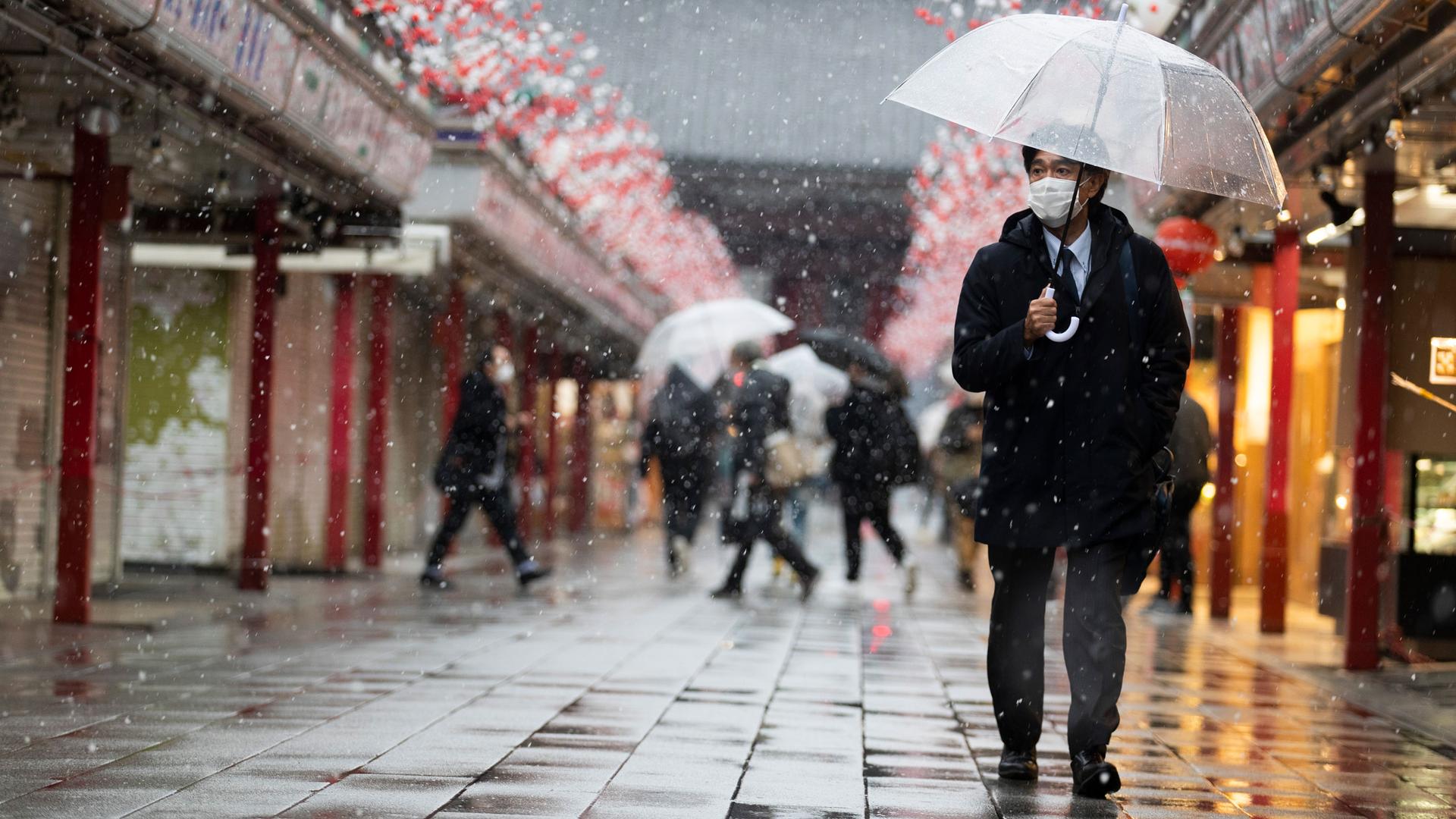 A man is shown carrying a clear umbrella and wearing a face mask while walking with his left hand in his pocket.