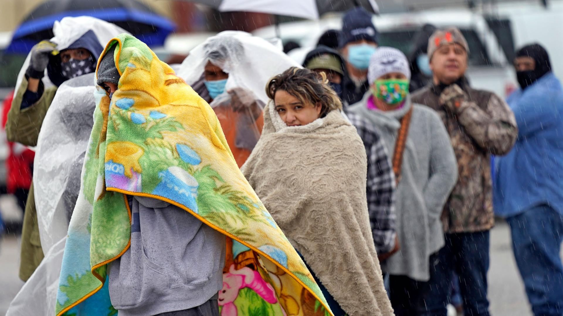 A line of people are shown with many people wrapping large blankets around themselves.