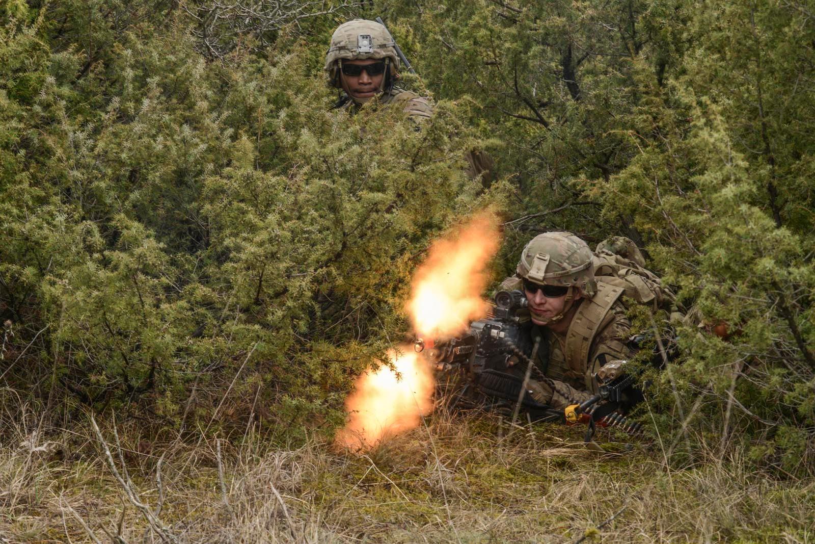 Two soldiers hunch down in a bush. The photo displays the bullets from one of the guns mid-fire.