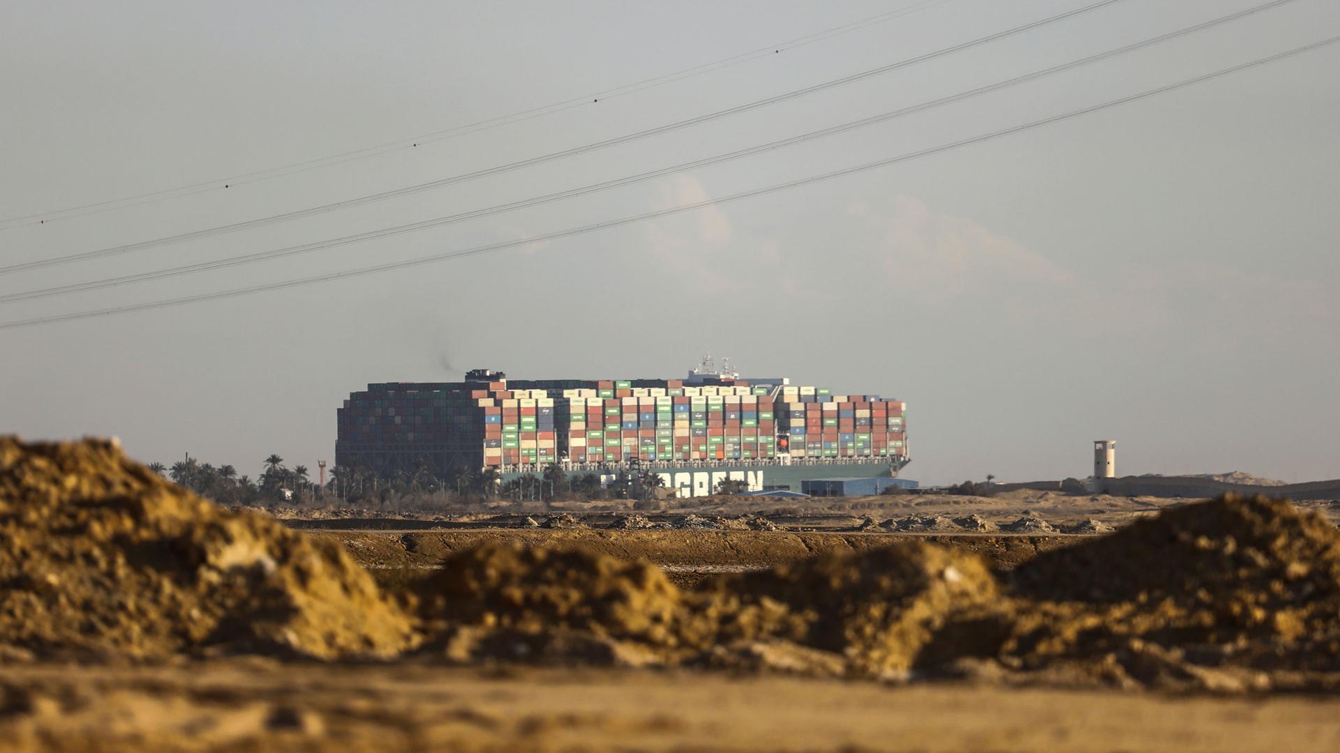 A large cargo ship is show in the distance towering over the sandy landscape with hundreds of containers stacked on top.