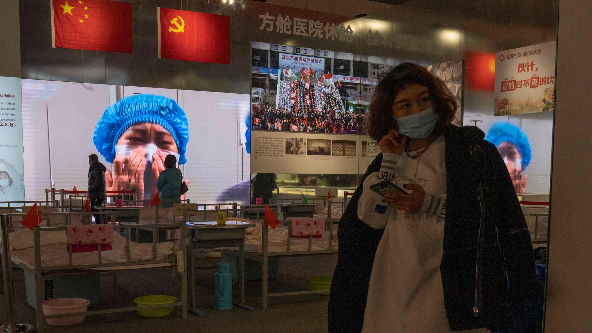 A woman is shown holding a mobile phone and wearing a face mask with an image of a medical worker on display in the background.