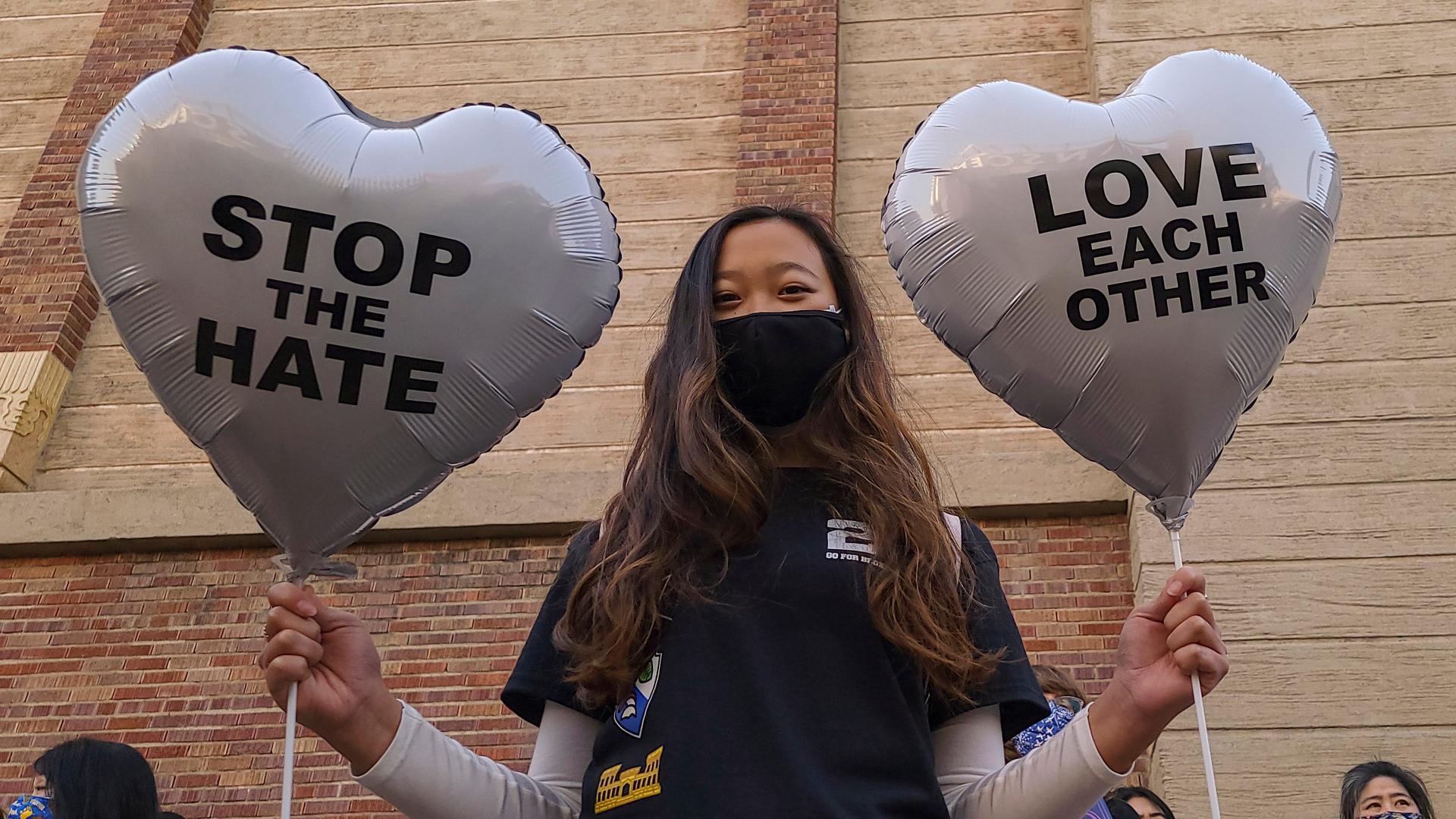 A young person holds up two balloons in heart-shapes that read Stop the Hate and Love Each Other