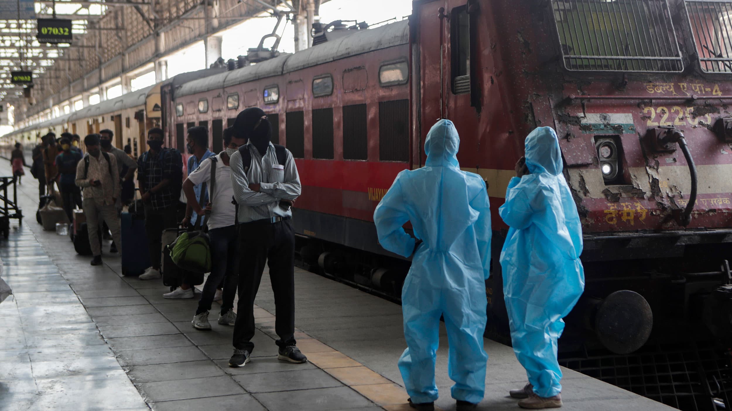 A long line of people are shown standing next to a train with two health care workers, shown at the end of the platform, wearing protective clothing.