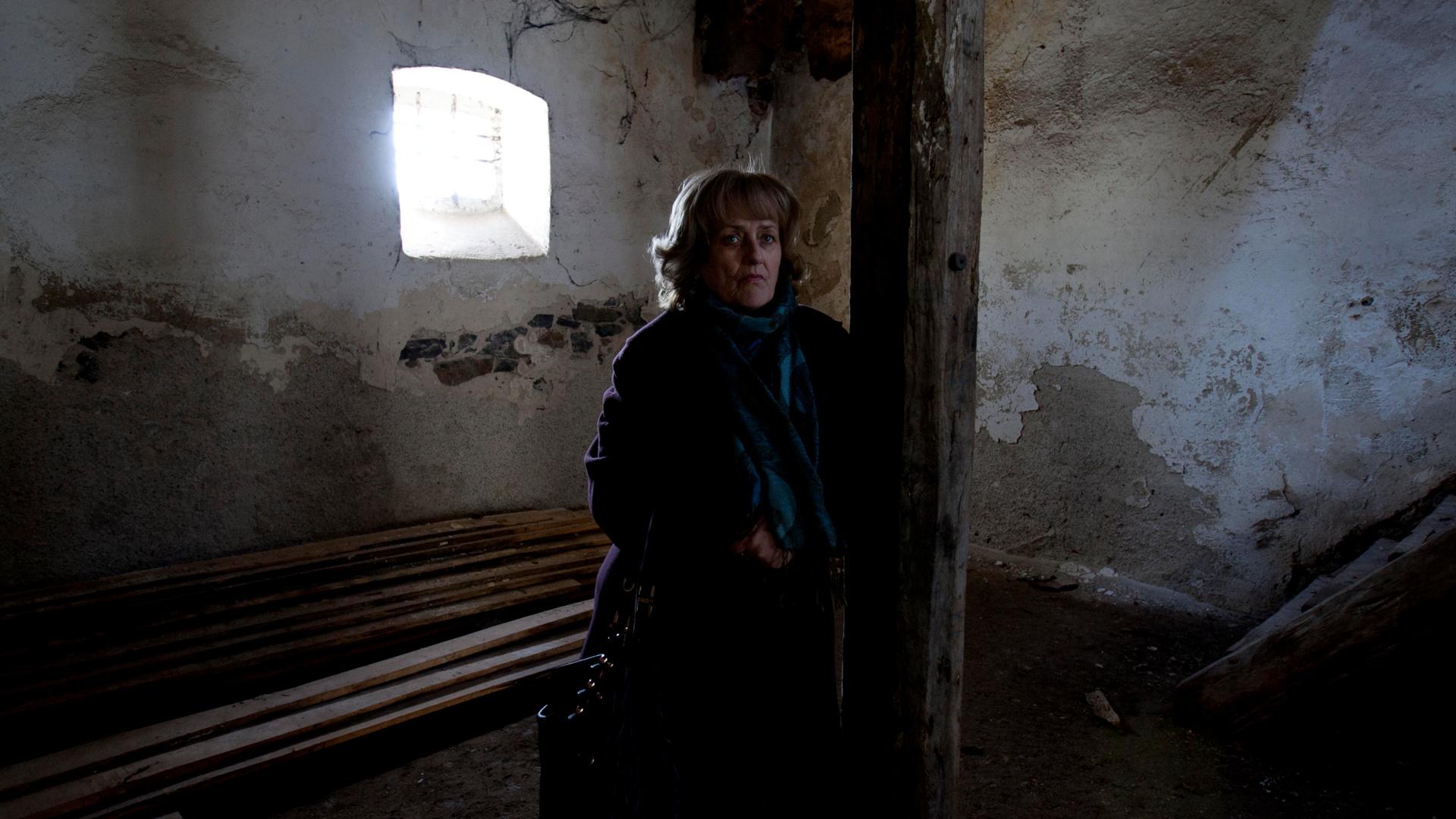 A woman stands in an abandoned building with light coming through the window.