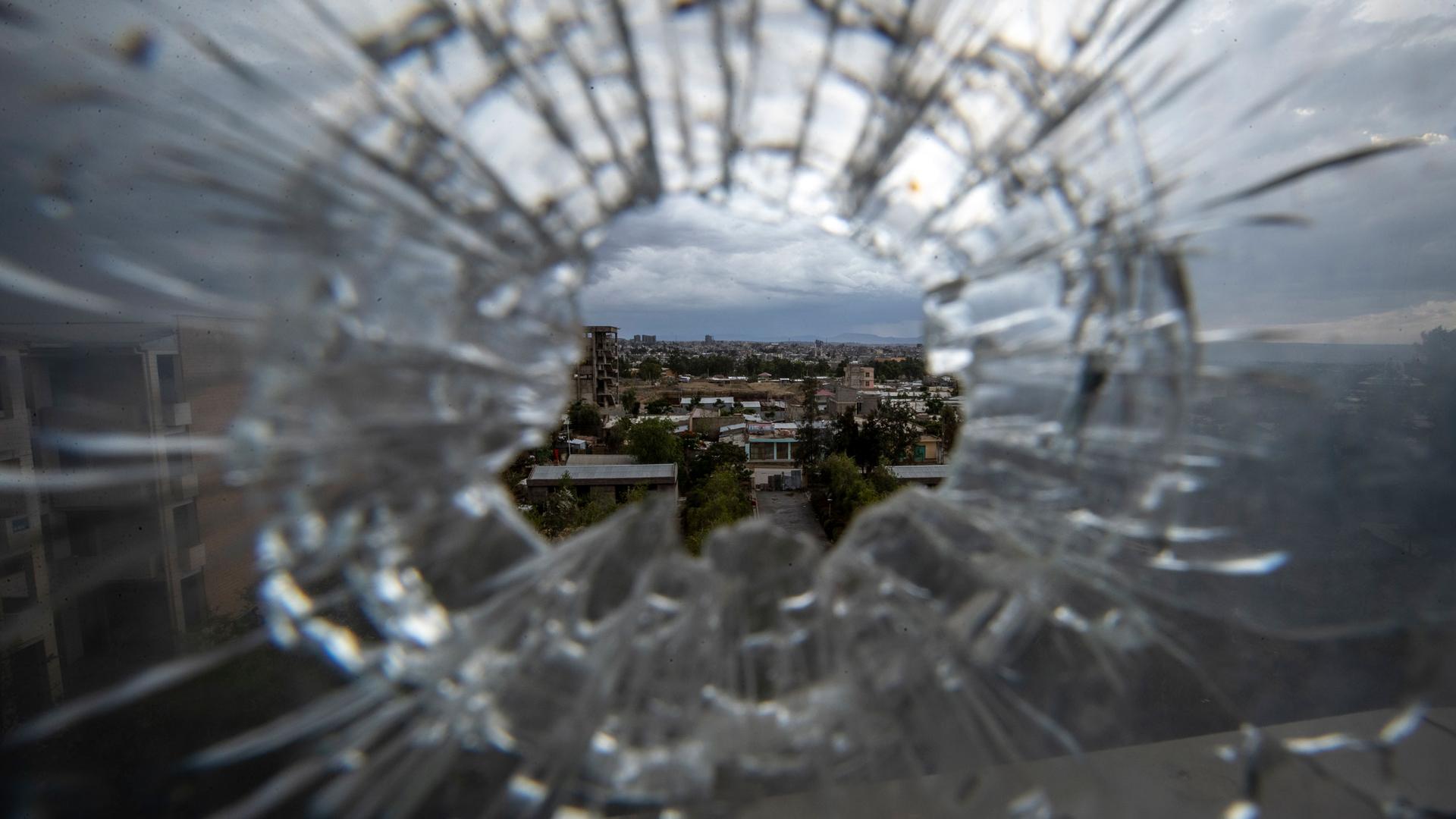 A circular hole is shown in a in a glass windown with cracks radiating from the center and through the hole buildings can be seen.